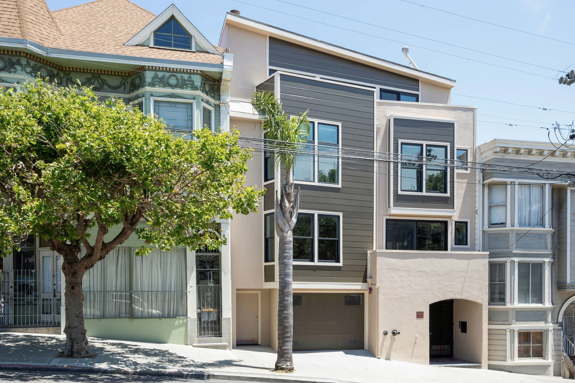 Modern multi-story urban home exterior in San Francisco featuring grey horizontal siding, tan stucco accents, and black-framed windows tucked between classic Victorian houses.
