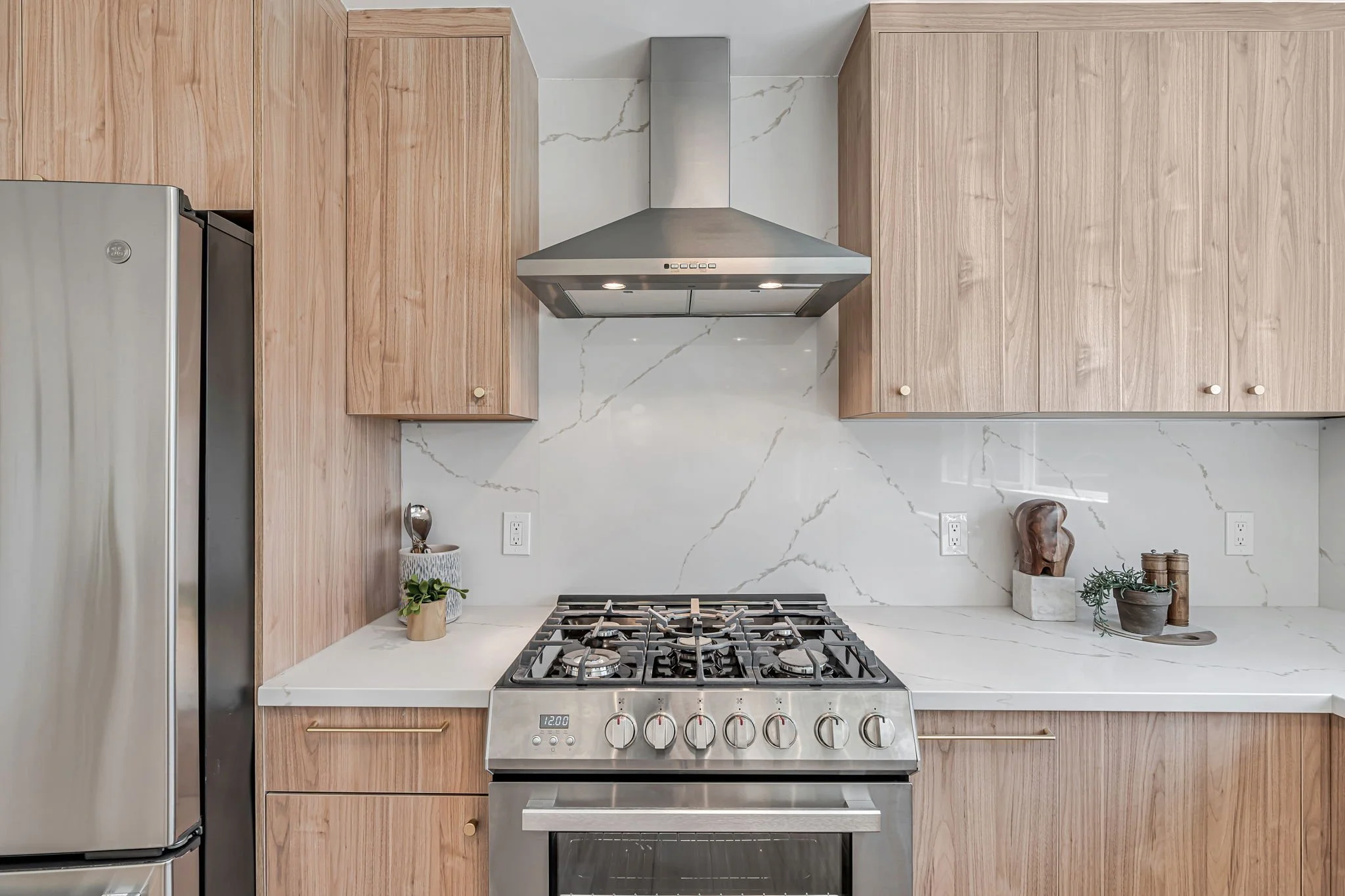 A straight-on view of the stainless steel five-burner gas range and oven. The stove is set against a matching white marble-look quartz backsplash and flanked by minimalist light wood cabinets with elegant gold pulls.
