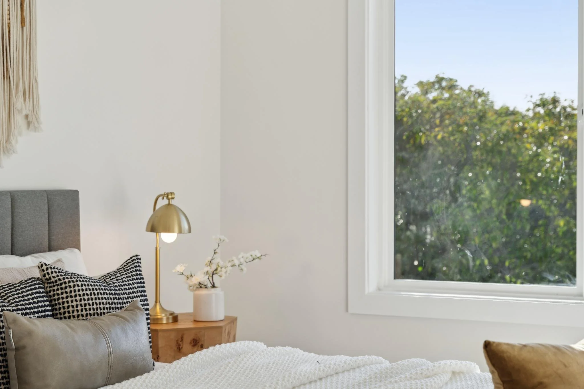 Close-up detail of a bedside setup in a luxury home with a burl wood nightstand, a modern gold lamp, and a view of lush green trees through the window.