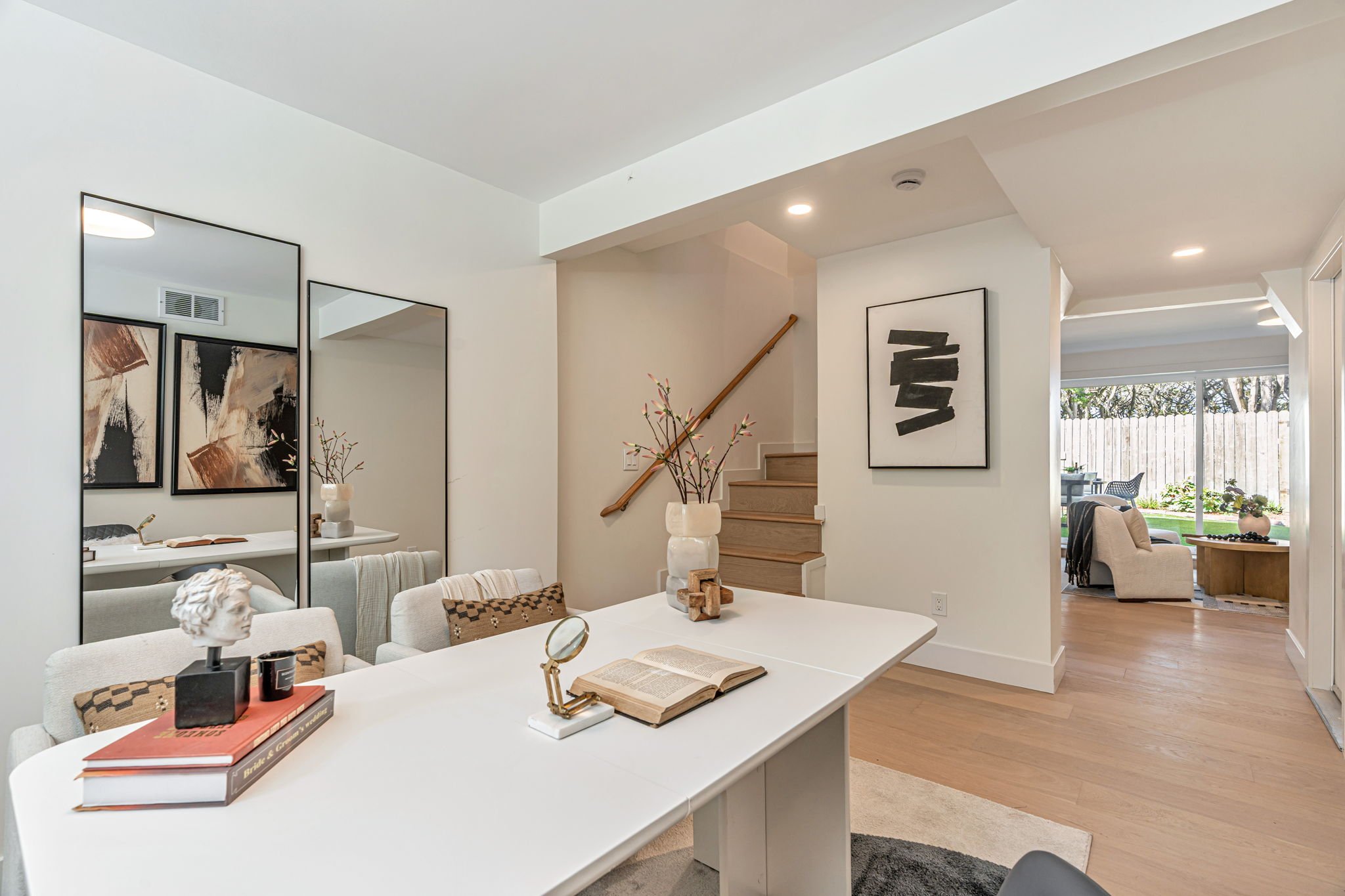 Modern workspace layout showing a white desk with gold accents, looking toward an open hallway that leads to a sunlit media room and backyard.