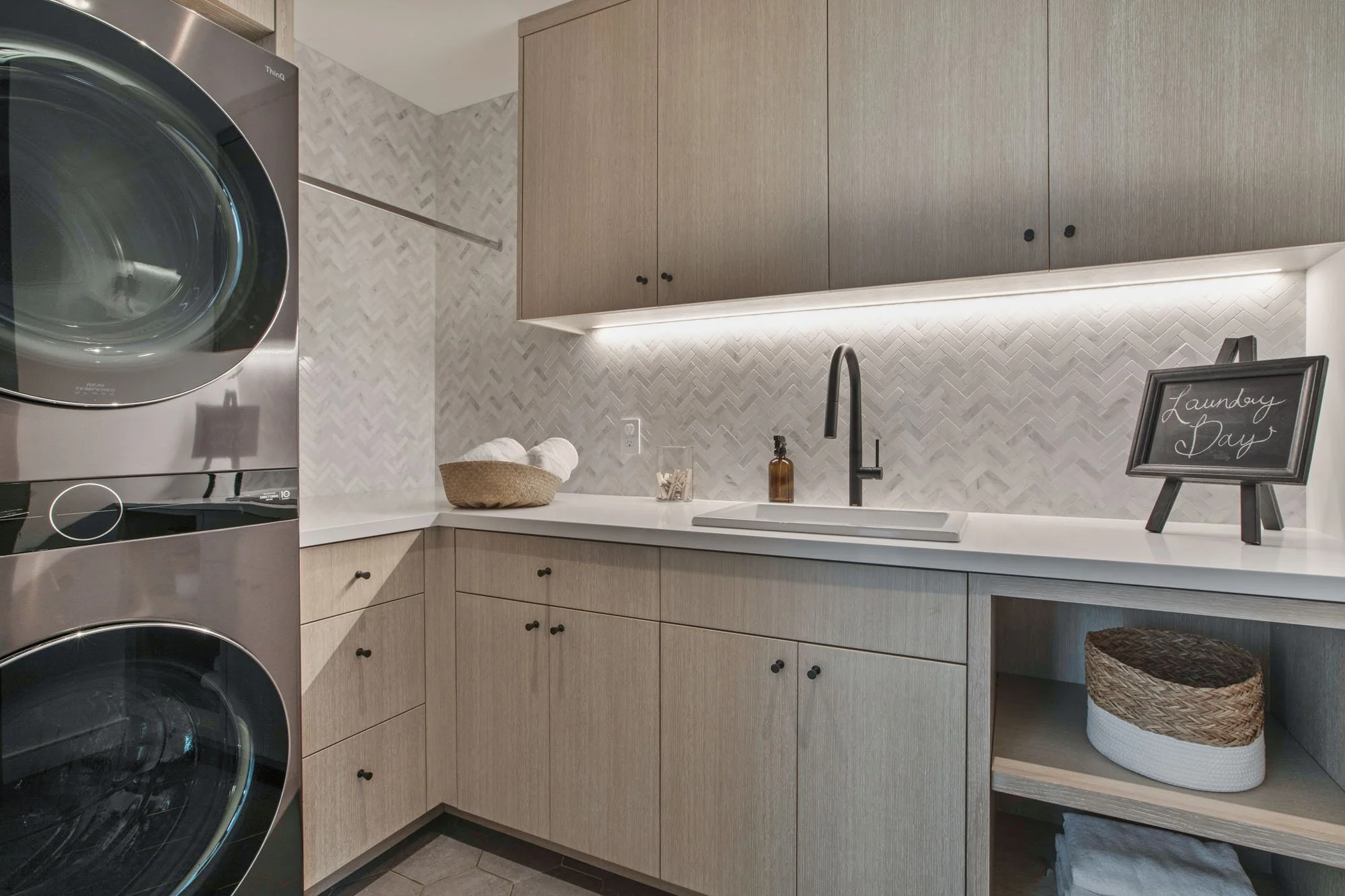 Luxury laundry room featuring LG ThinQ stackable washer and dryer, custom light oak cabinetry, and a white marble herringbone tile backsplash.