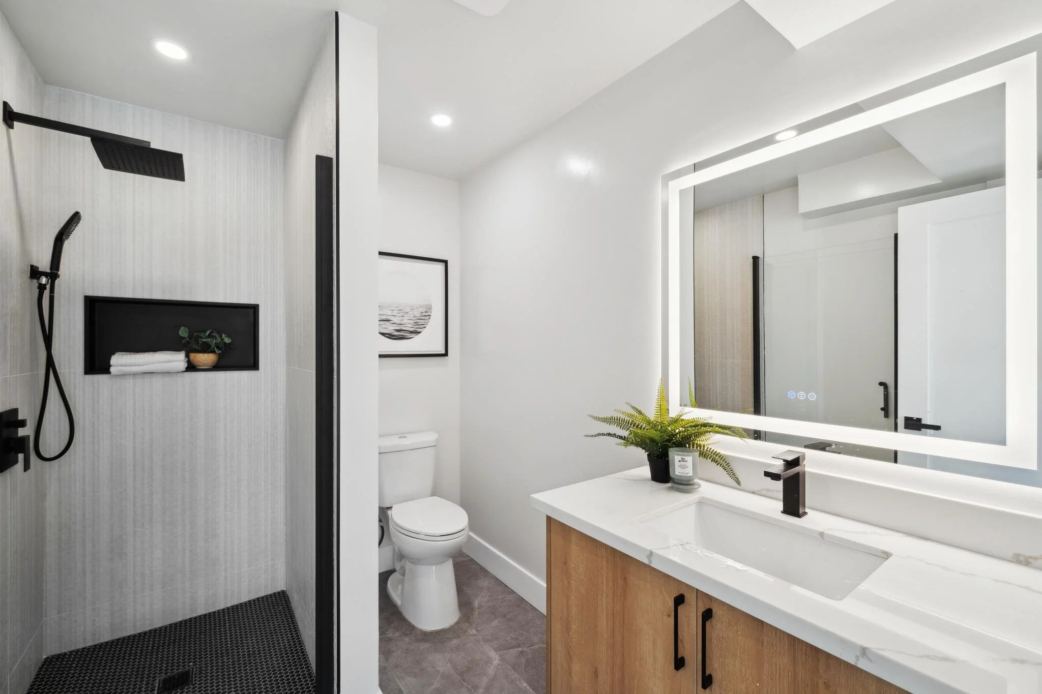 Renovated guest bathroom with a wood vanity, white quartz countertop, back-lit LED mirror, and a walk-in shower with vertical stacked tiles.