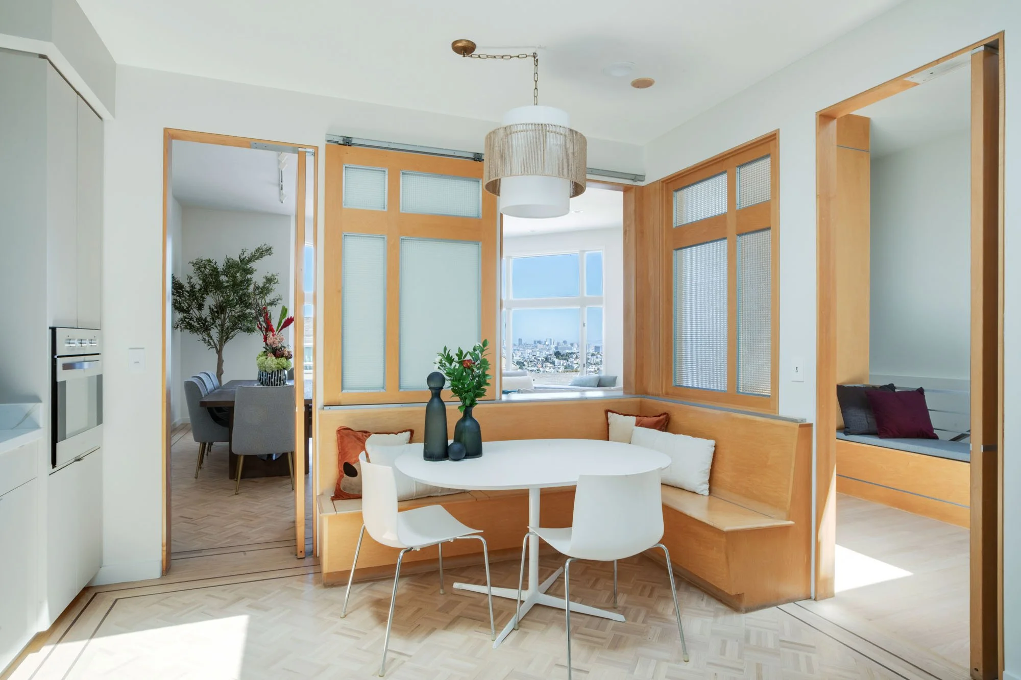 Modern San Francisco kitchen breakfast nook featuring custom light-wood built-in bench seating, parquet floors, and a pass-through window with a panoramic view of the downtown skyline.