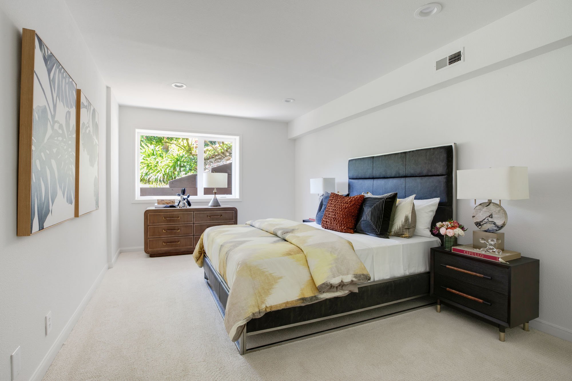A bright, modern San Francisco bedroom featuring a dark leather tufted headboard, geometric yellow and white bedding, and a fluted wood dresser under a large window overlooking a lush garden.