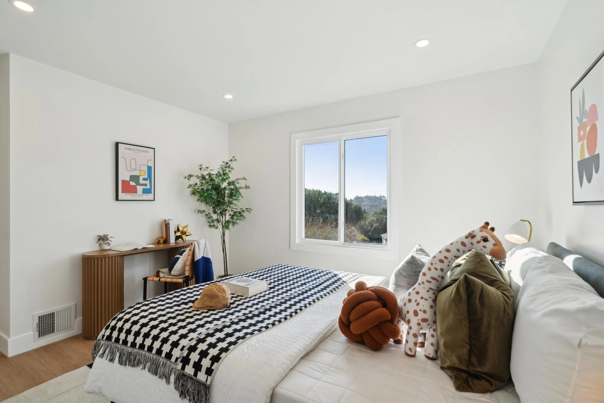 Bright second bedroom with minimalist decor, featuring a large window with views of San Francisco’s greenery and a playful giraffe plush for a nursery-style feel.