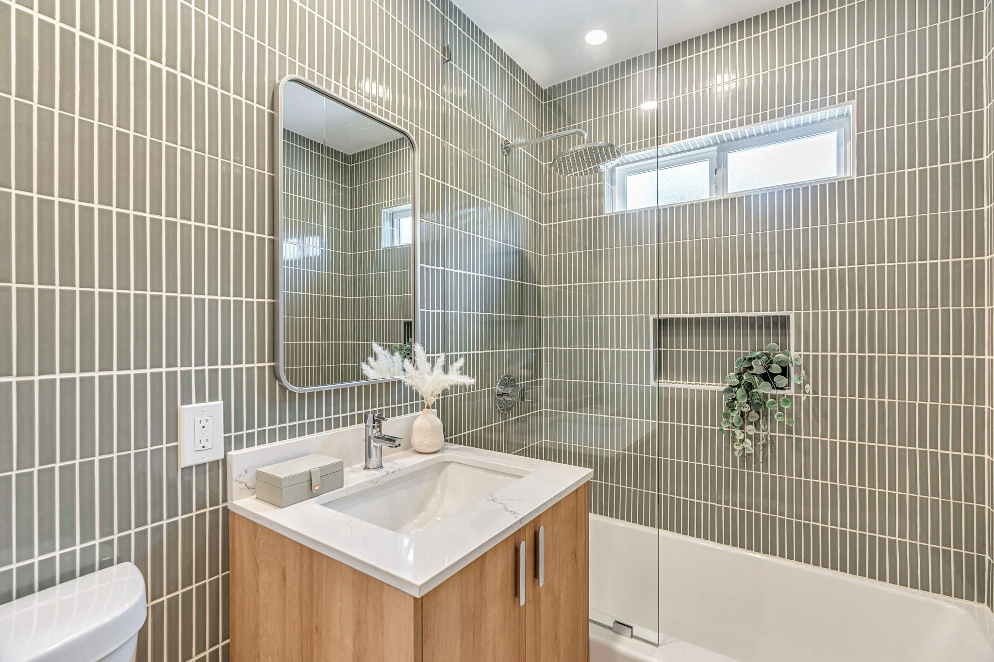 A modern ensuite bathroom featuring soft sage green vertical stacked tiles. It includes a light wood vanity with a white marble top, a rectangular mirror, and a shower-tub combination behind a glass partition.