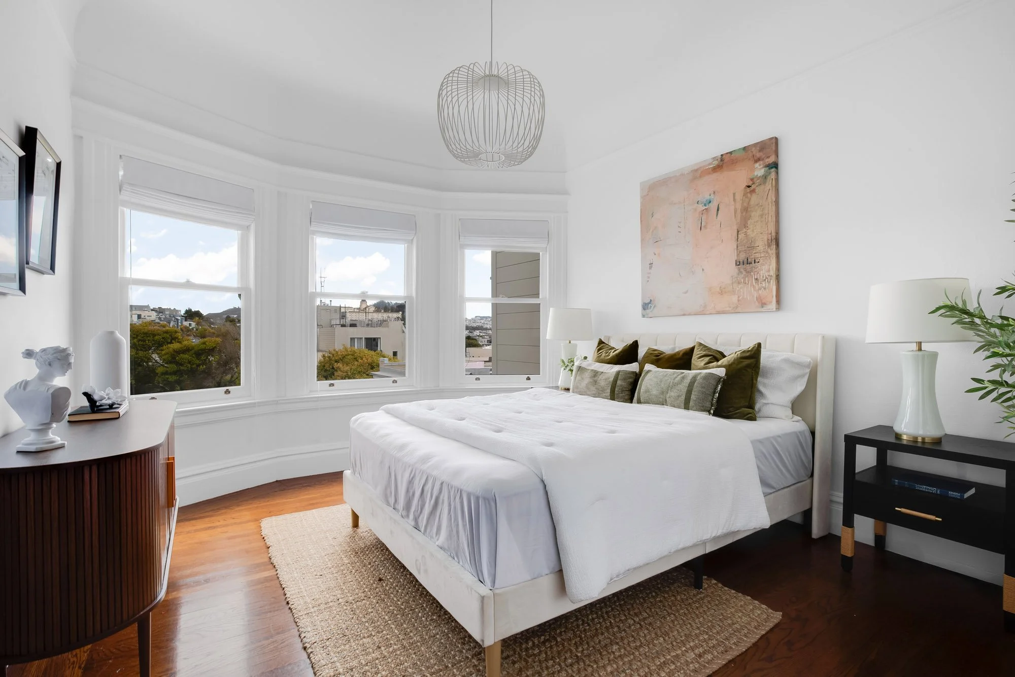 Light-filled primary en-suite bedroom with original curved bay windows, hardwood floors, and sweeping southern views over Presidio Heights rooftops and treetops, San Francisco luxury Victorian flat