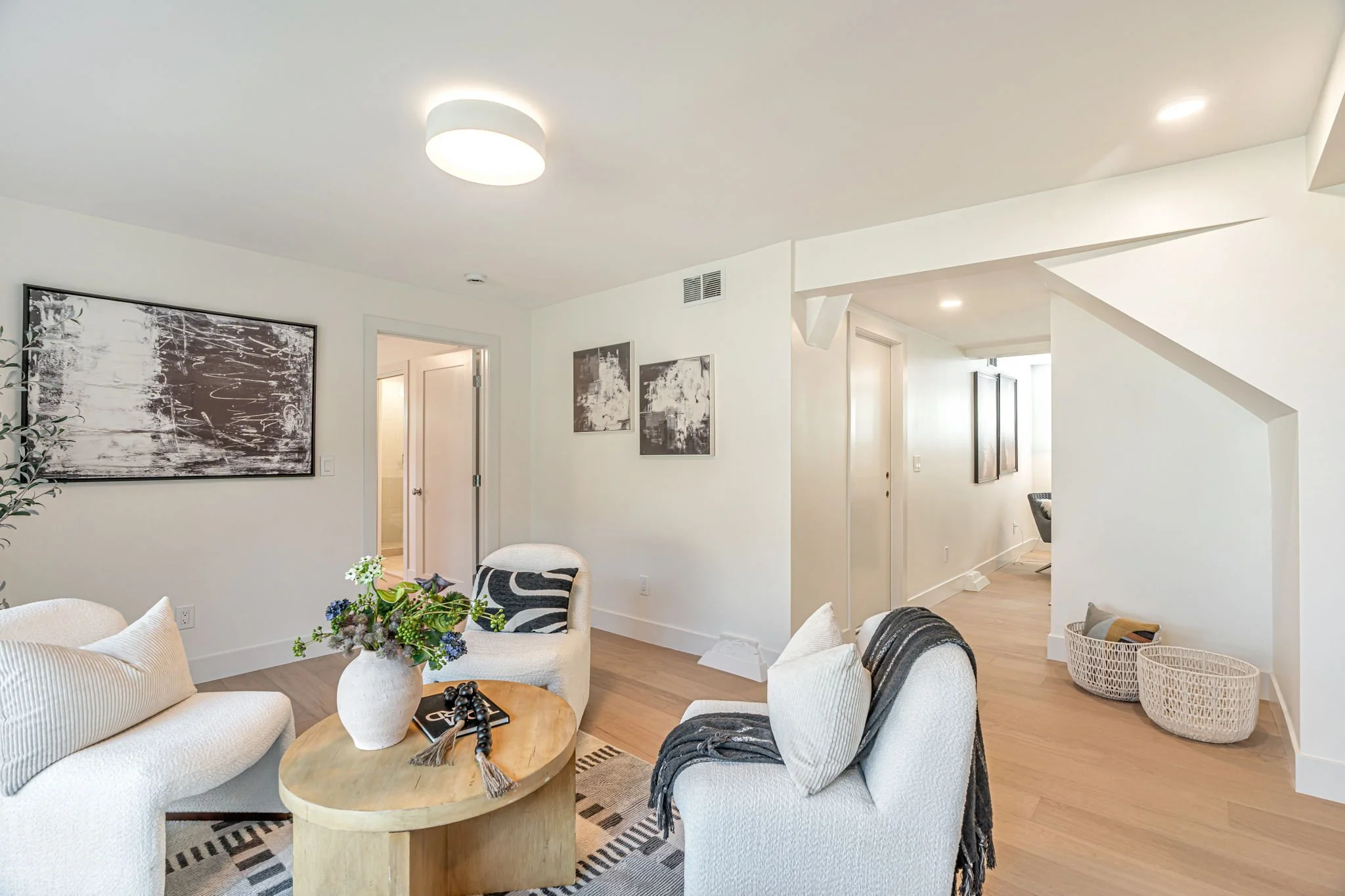 Modern residential media room layout featuring white bouclé chairs, a round oak coffee table, and abstract wall art, highlighting a seamless transition to the home office and hallway.