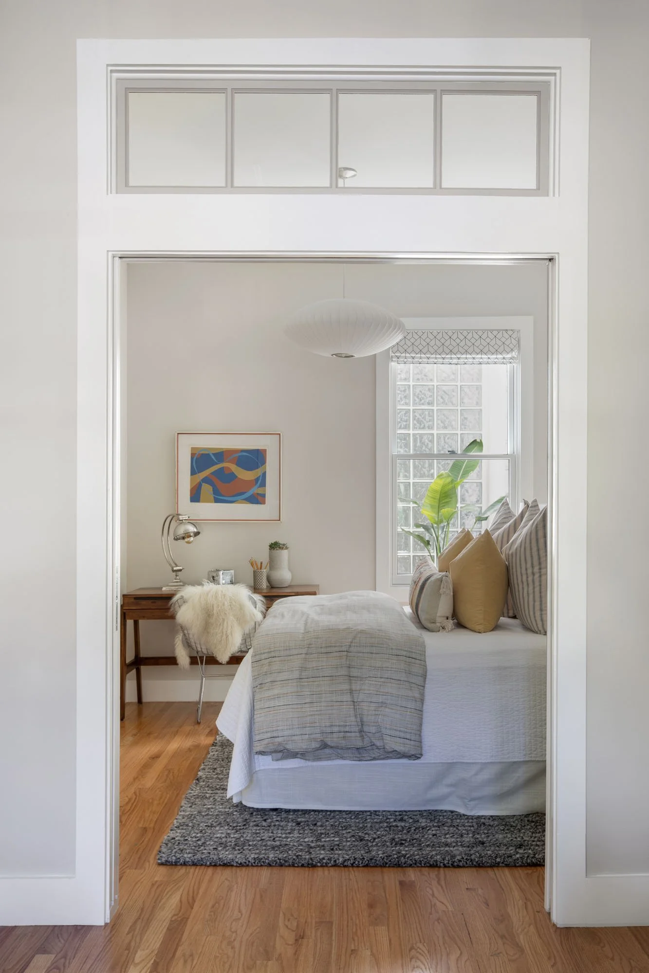 View into a cozy bedroom featuring a wooden writing desk with a white faux fur chair, a neutral bed, and a large window with garden views.