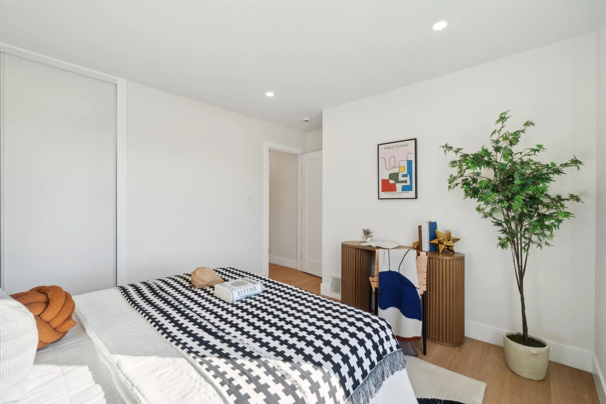 Interior view of a guest bedroom showing a built-in closet, light oak flooring, and a stylish fluted wood desk under an abstract Pablo Picasso art print.
