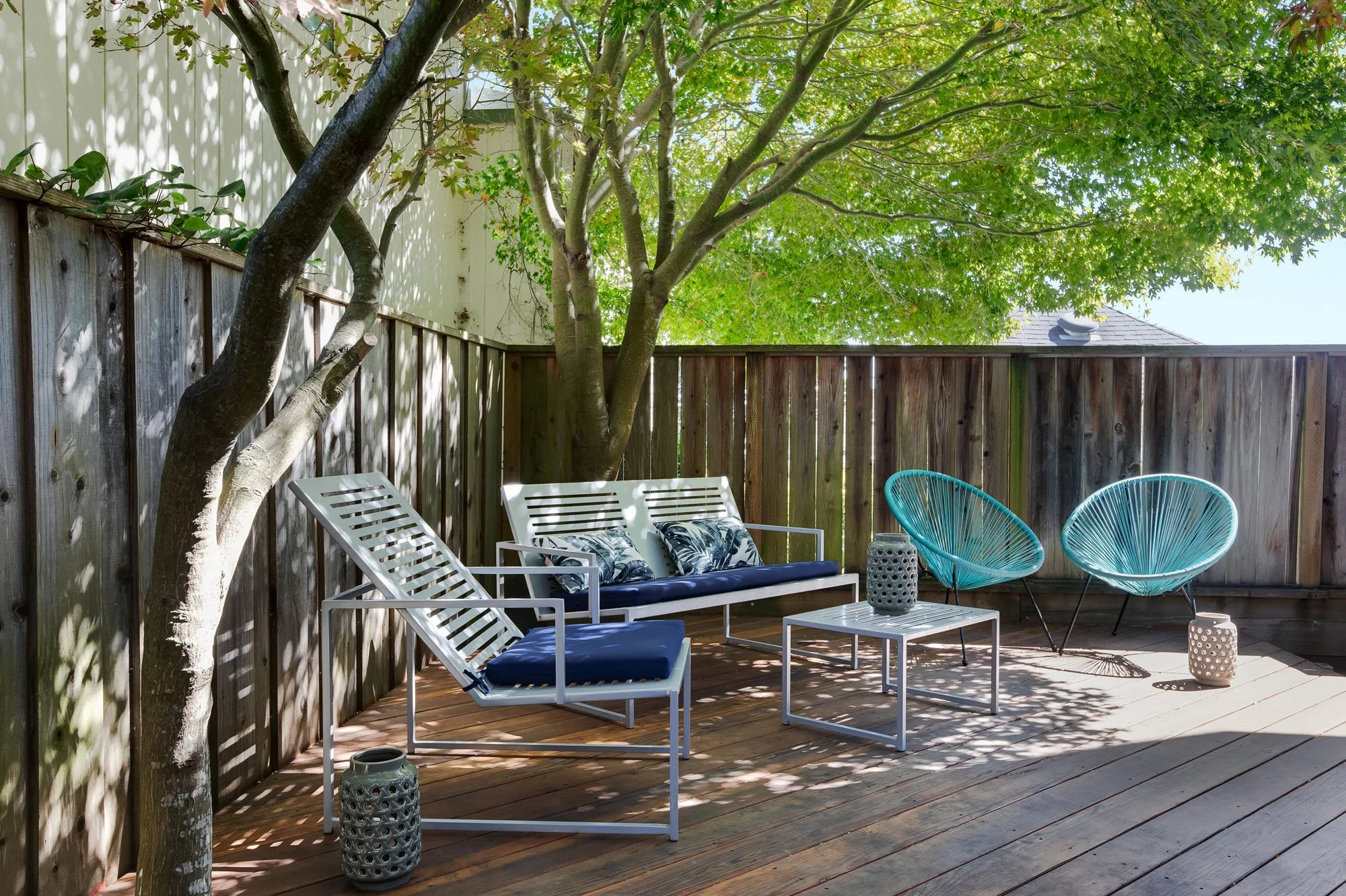 A private wooden sun deck in a secluded backyard, featuring modern white outdoor furniture, turquoise Acapulco chairs, and a mature Japanese Maple tree.