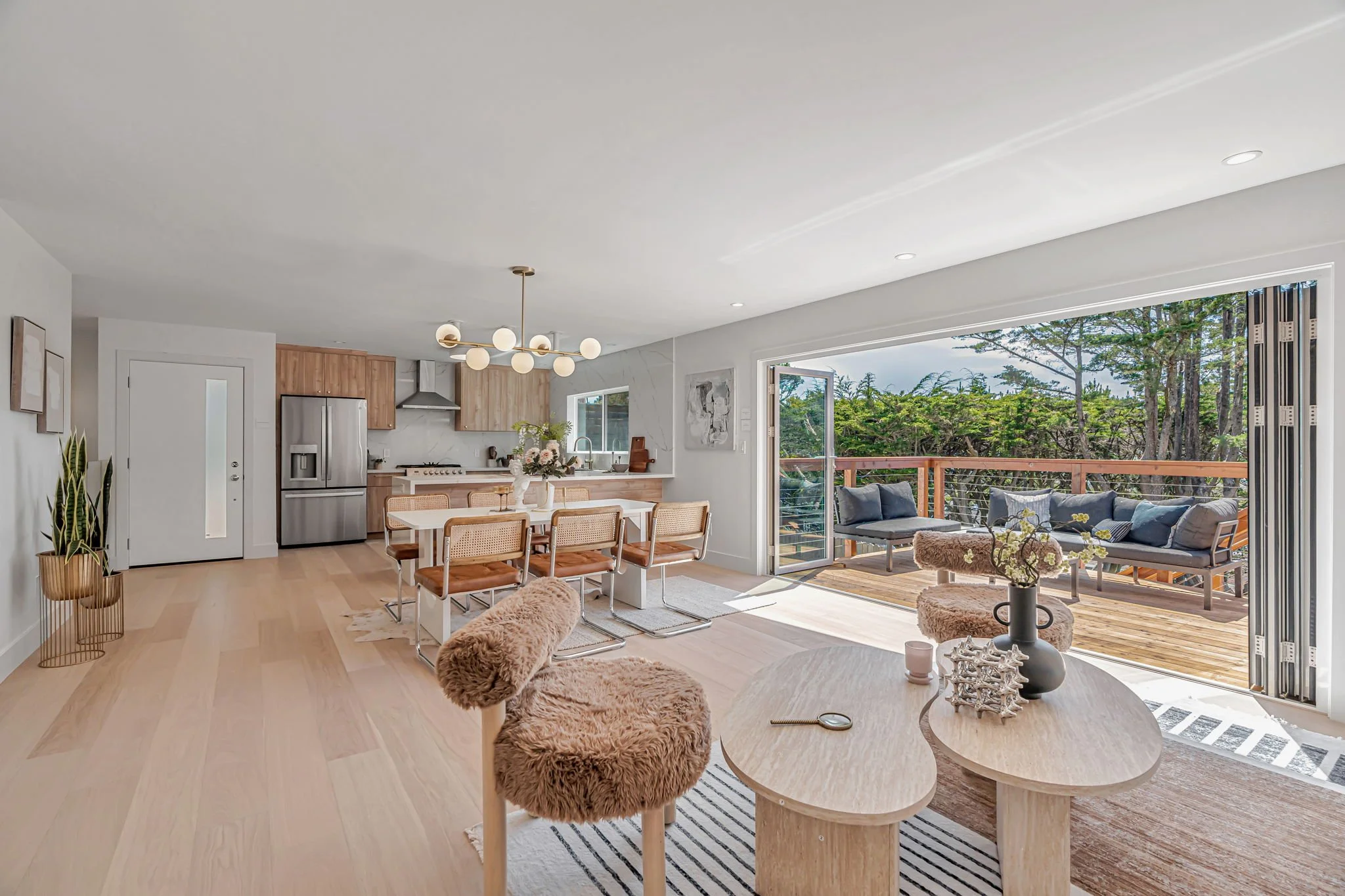 A wide view of the interior showing the kitchen, dining area, and living room. A massive accordion glass door is fully retracted, seamlessly connecting the indoor living space to a large wooden deck surrounded by trees.