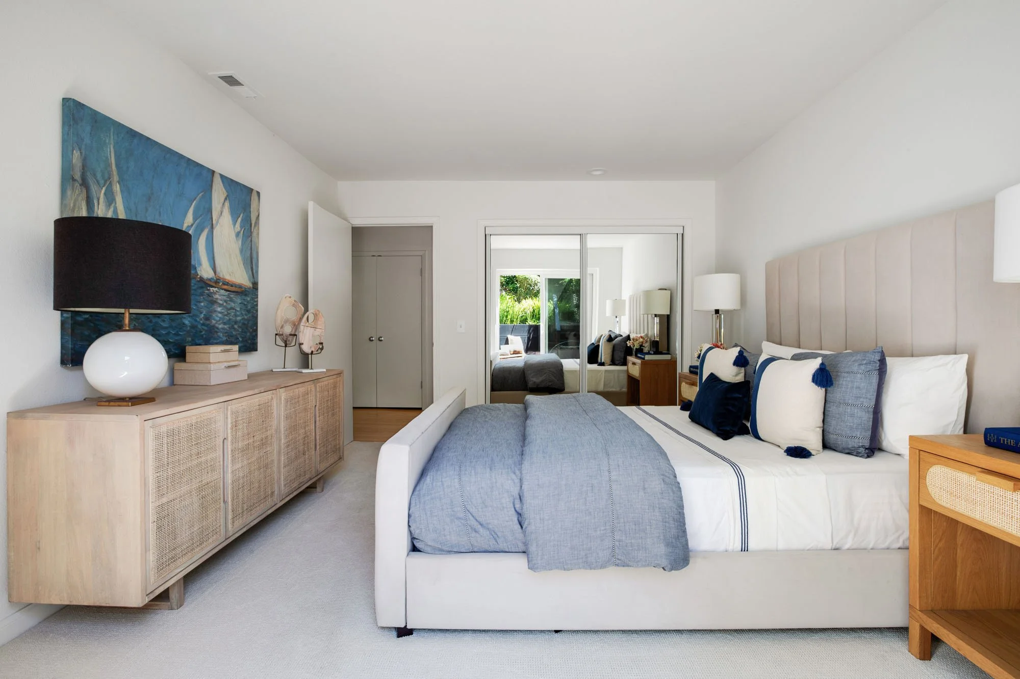A spacious bedroom featuring a large sailboat oil painting, a long wood and cane sideboard, and mirrored sliding closet doors that reflect the natural light from the outdoor patio.
