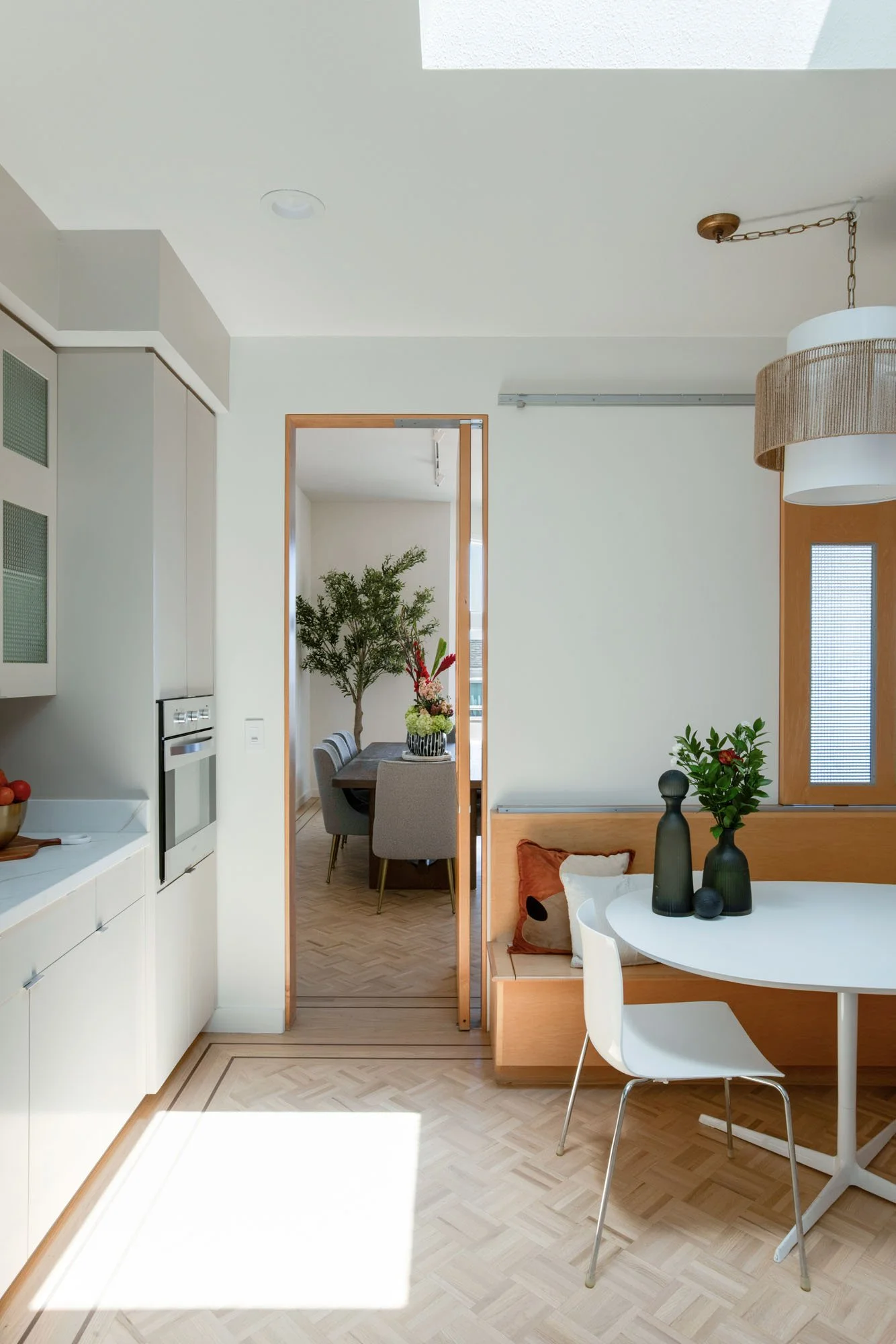 Bright kitchen interior showing a modern built-in oven and parquet hardwood floors, with an open doorway leading into a sun-drenched formal dining area.