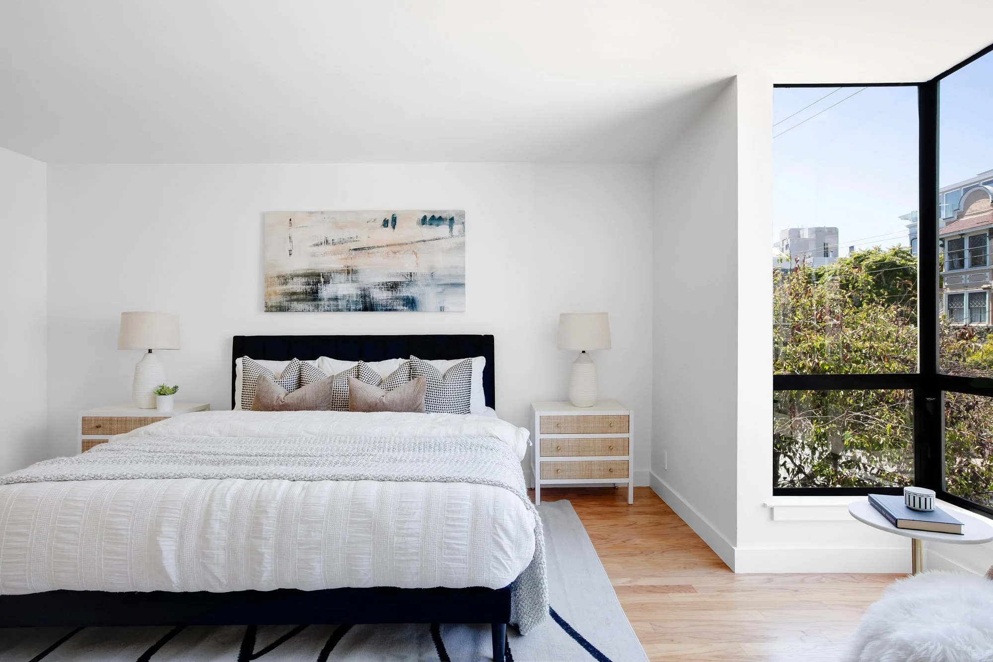 Primary bedroom at 932 Broderick Street #2 with a navy upholstered bed, large corner windows framing leafy neighborhood views, and rattan nightstands.