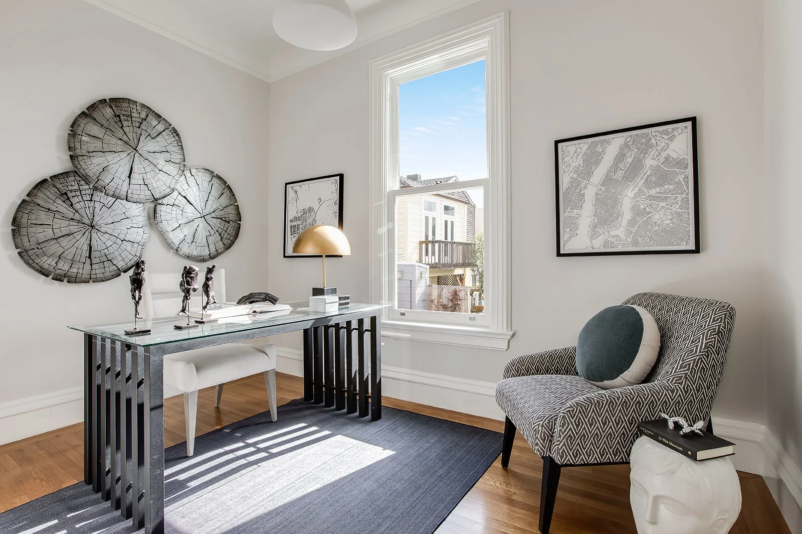 Sun-drenched luxury home office in a renovated Pacific Heights Victorian, featuring a designer glass-top desk, sculptural wall art, and a cozy reading nook with scenic neighborhood views.
