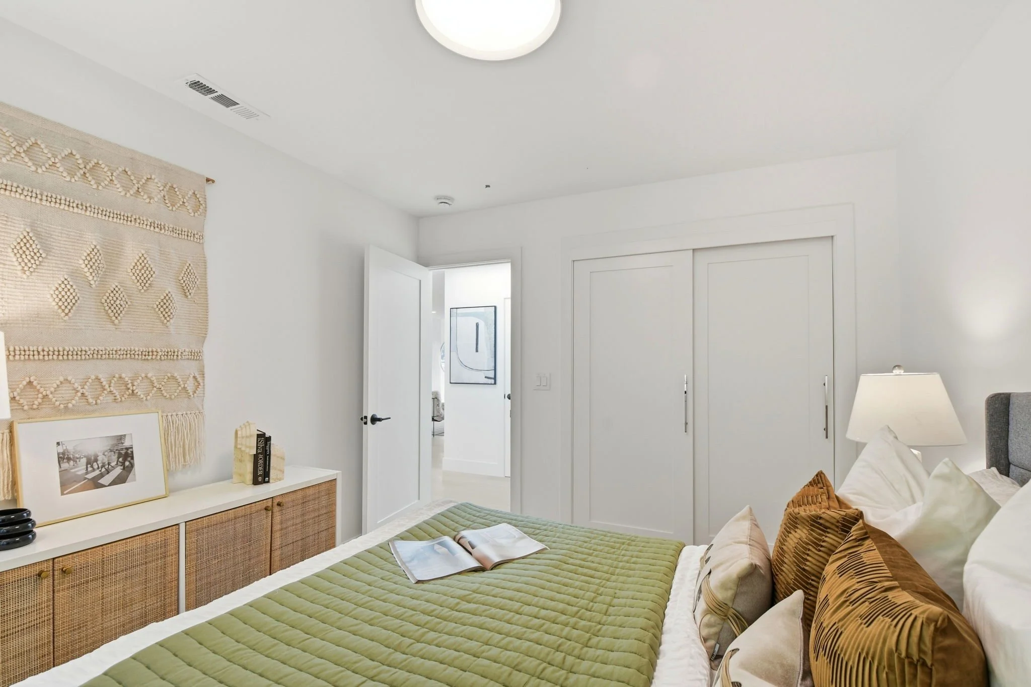 Light-filled guest bedroom with a sage green quilt, woven boho wall hanging, and clean white architectural lines.