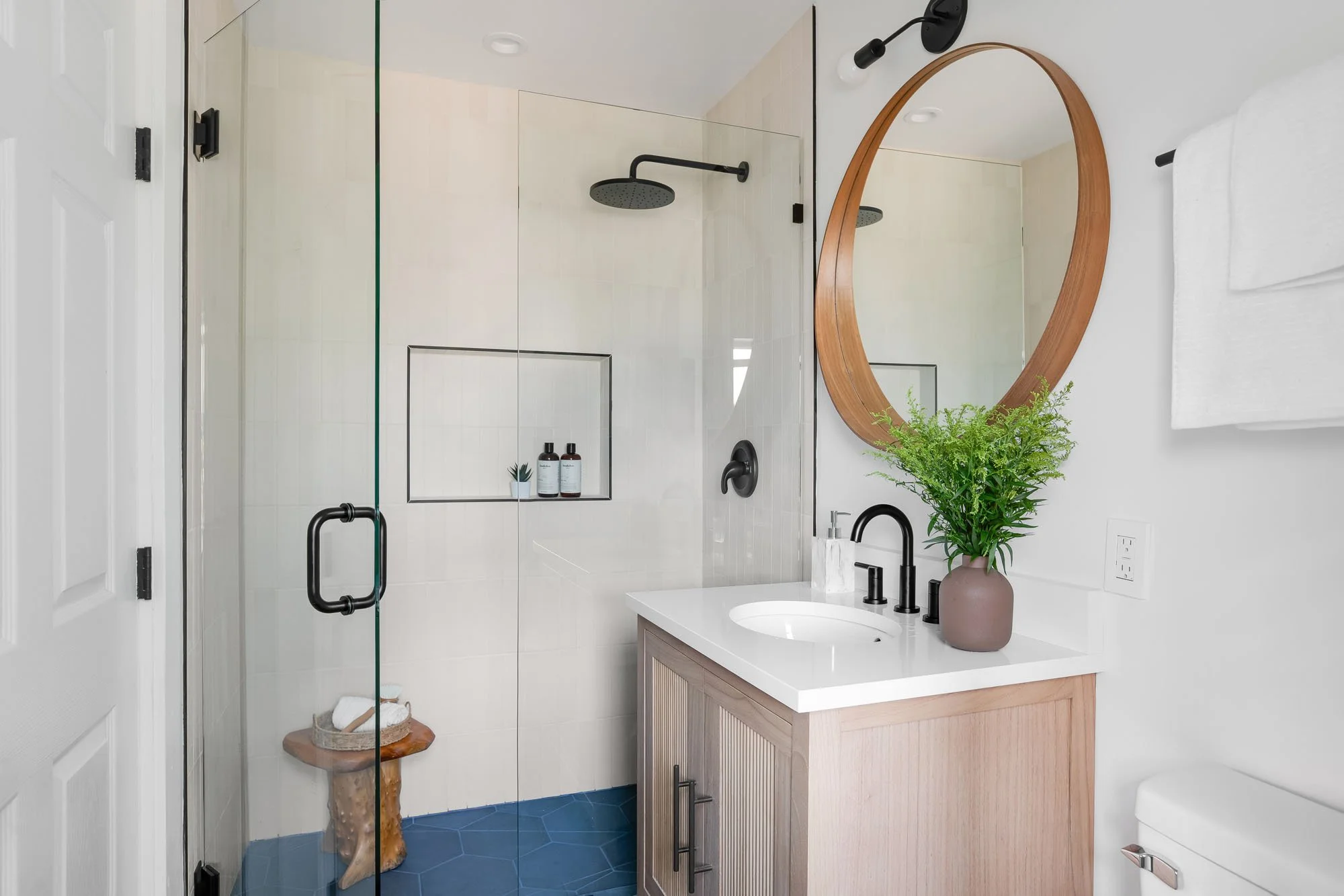 Modern bathroom featuring a walk-in glass shower with white vertical tiles, a wooden vanity with black hardware, and blue hexagonal floor tiles.