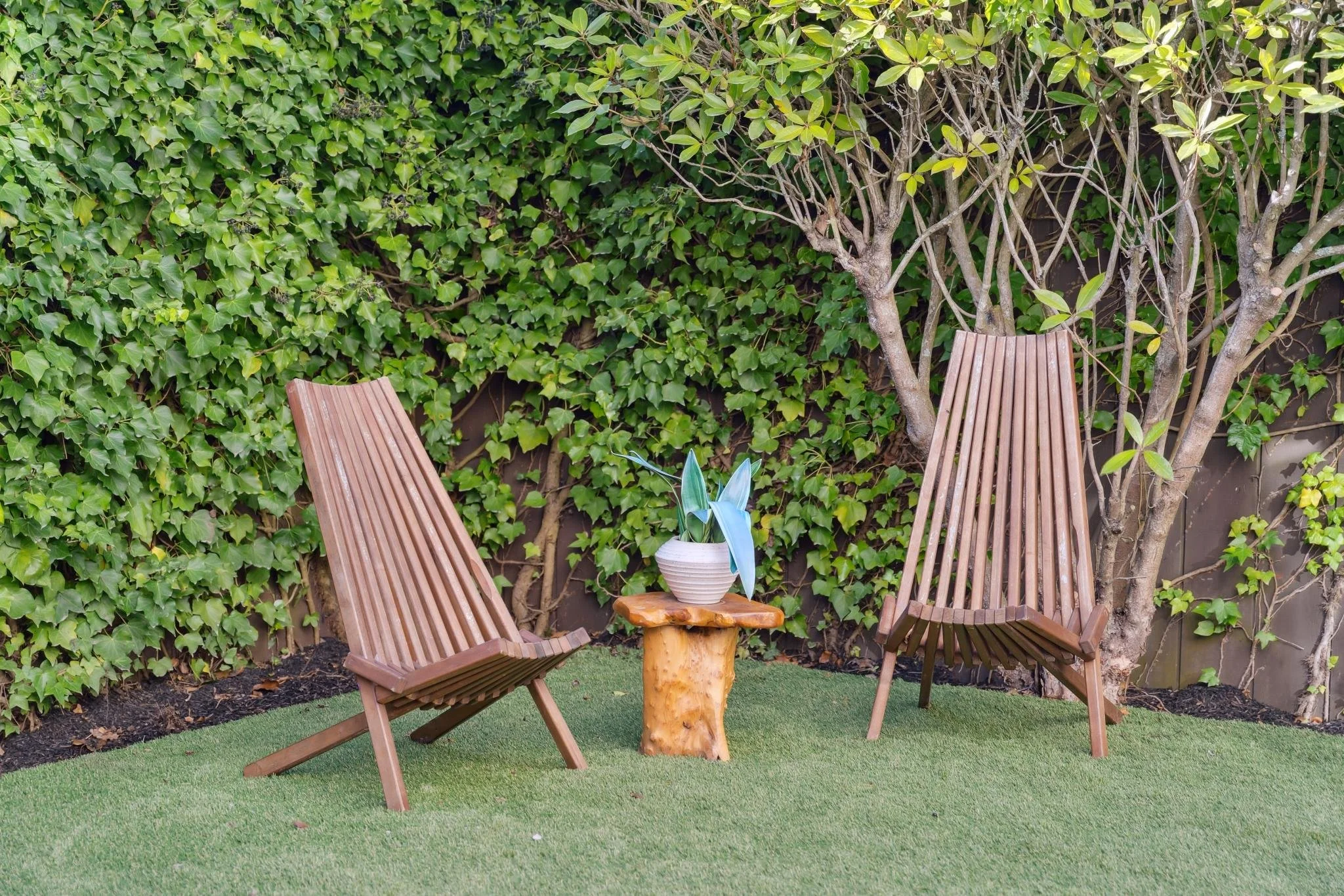 Detail of a cozy backyard nook with mid-century style wooden chairs and a natural wood side table against a green ivy wall.