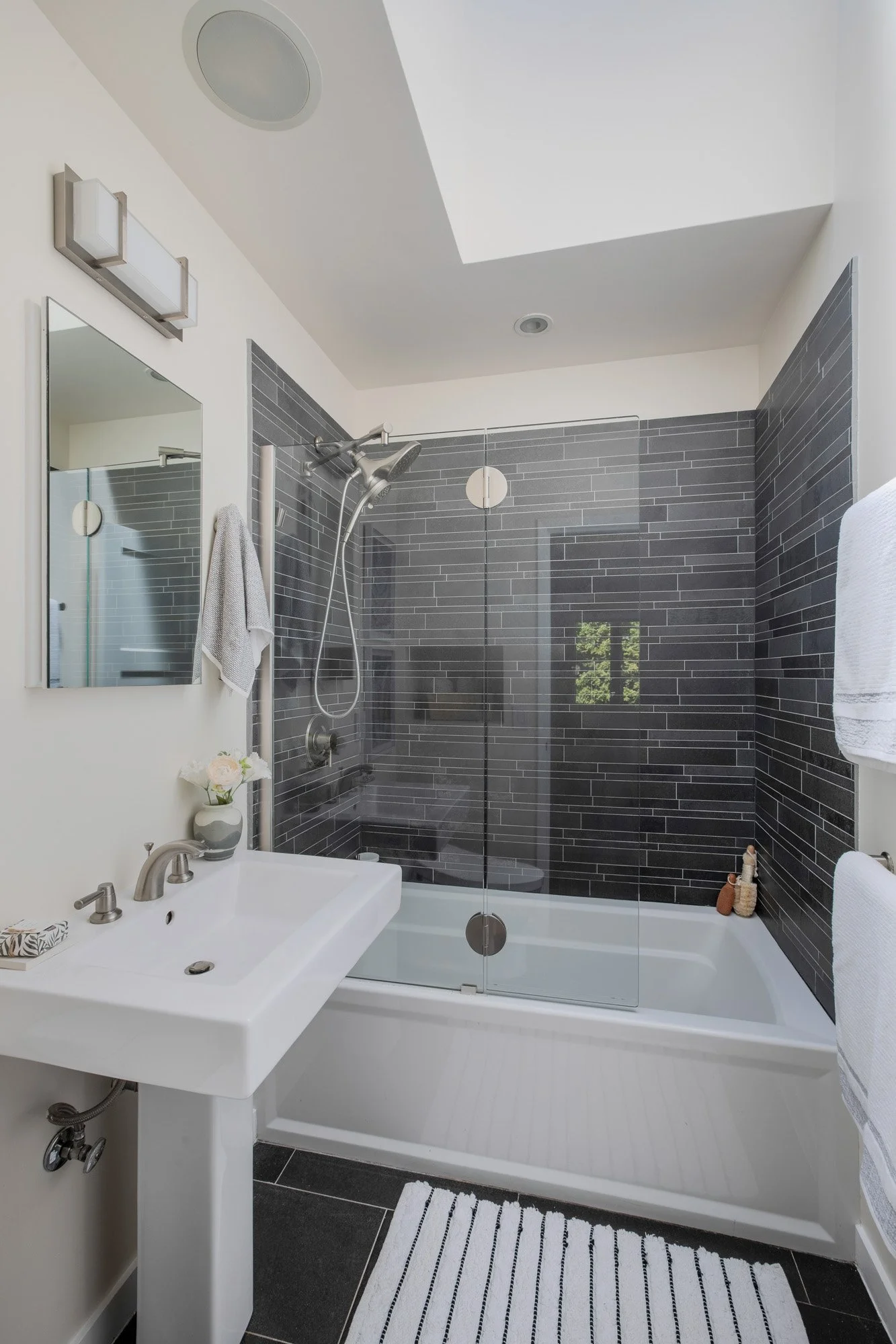Modern bathroom featuring a white pedestal sink and a glass-enclosed bathtub with charcoal gray subway tile.
