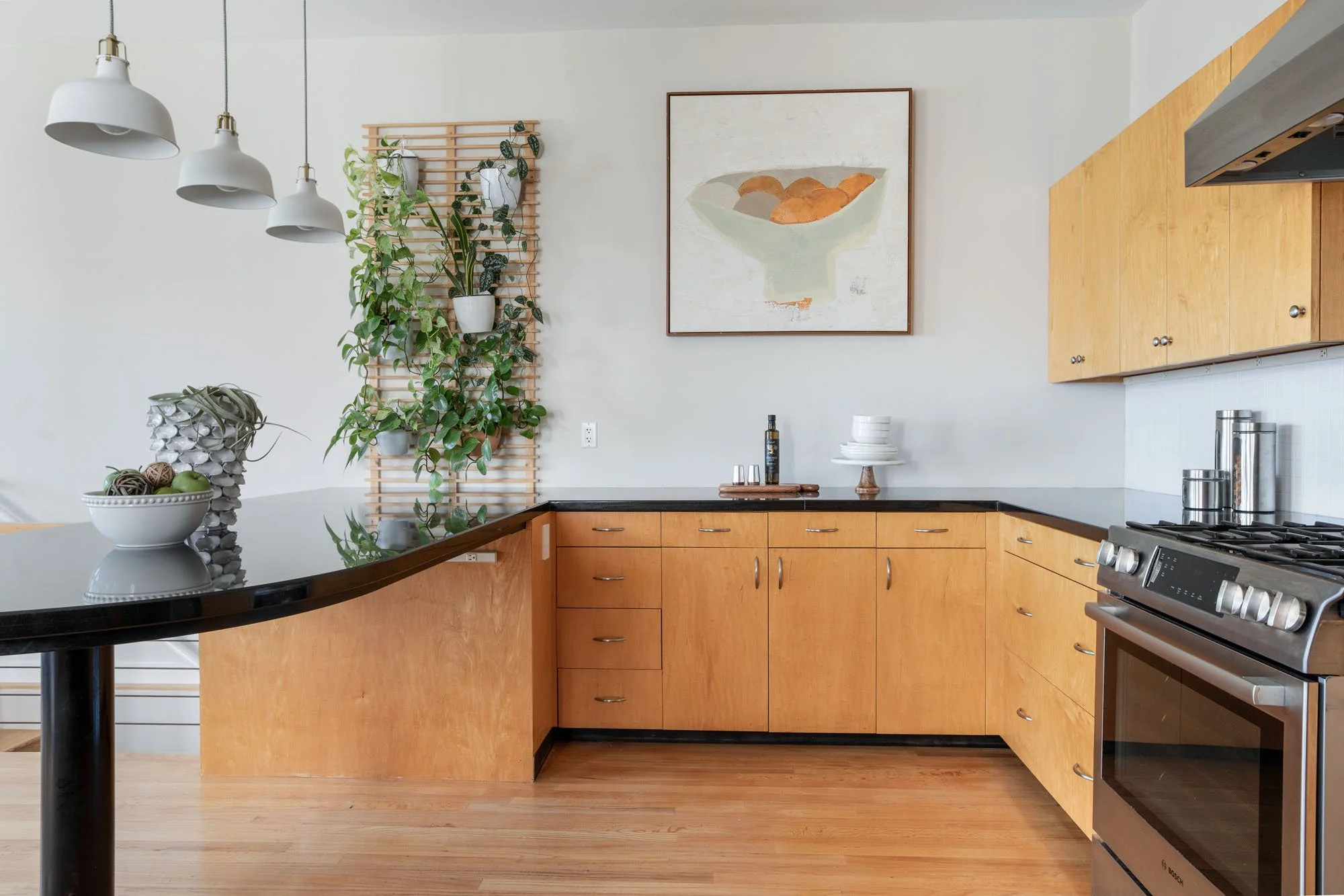 Modern U-shaped kitchen featuring light wood cabinets, black quartz countertops, and a stainless steel gas range with a unique vertical wall planter and abstract art.