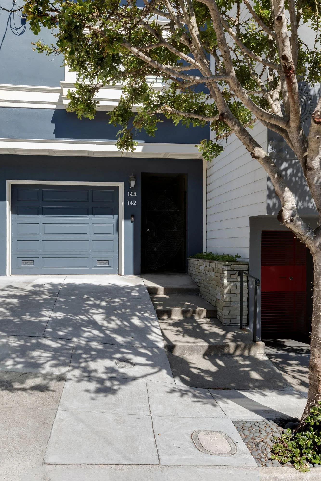Street-level view of the property entrance showing the "144-142" address numbering, a concrete walkway, a stone planter box, and a mature street tree.
