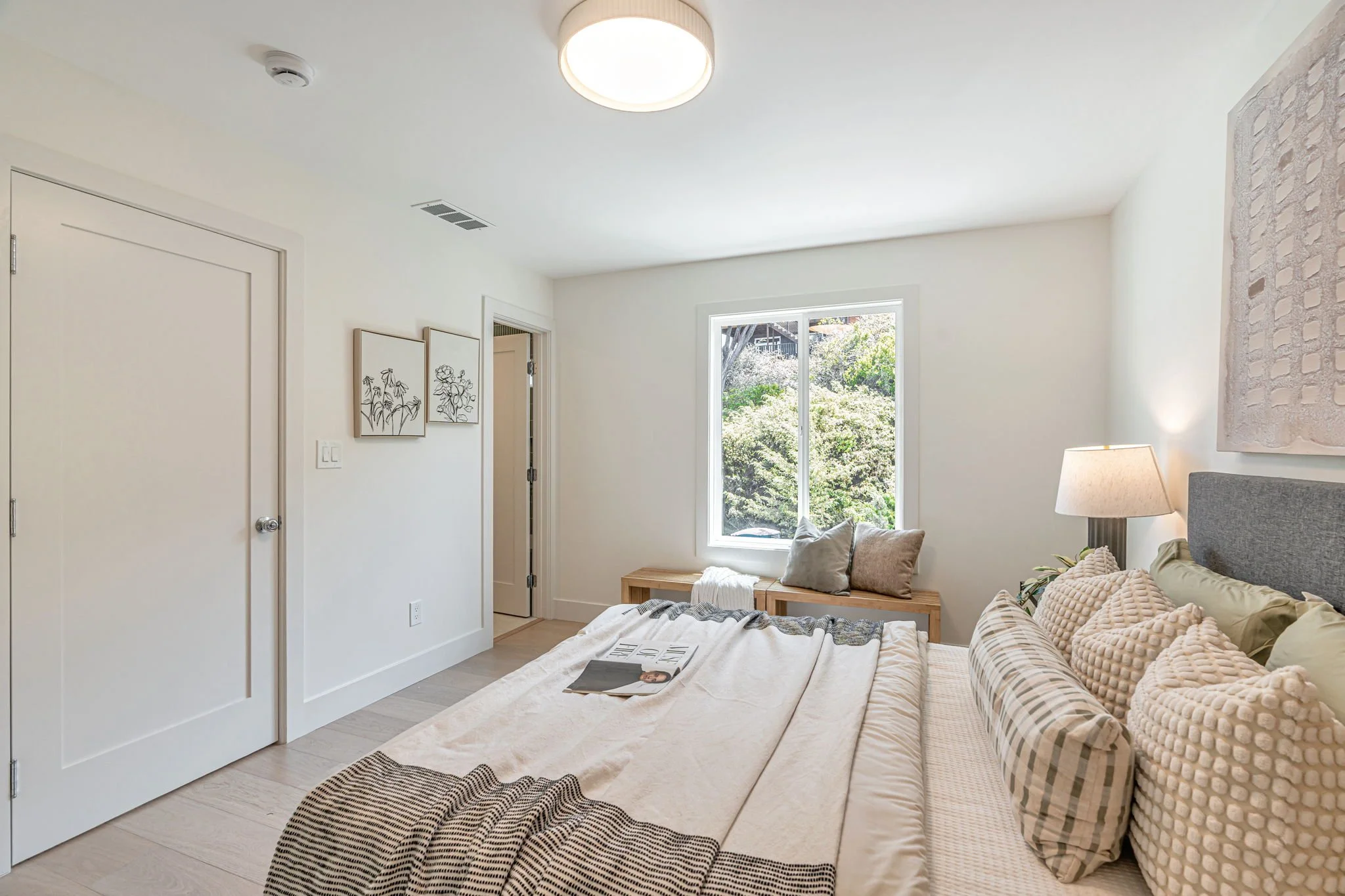 A perspective shot of the primary bedroom showing the clean, white interior and light wood flooring. A minimalist wooden bench sits beneath the large window, and the bed is styled with neutral-toned linens and textured throw pillows.