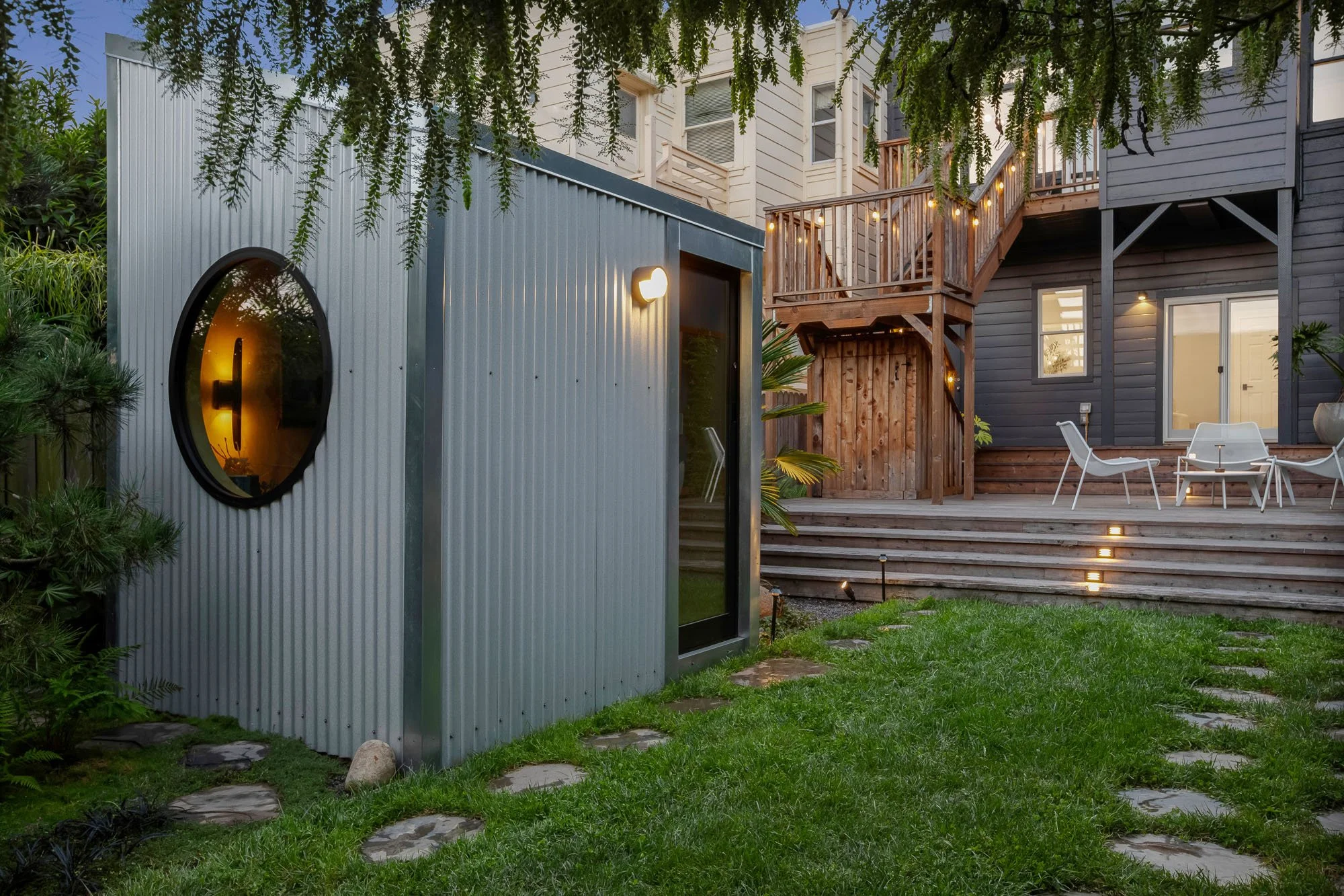 Twilight view of a modern backyard at 246 Detroit Street, SF, featuring an illuminated corrugated metal garden office, a multi-level wooden deck with string lights, and professional landscape lighting.