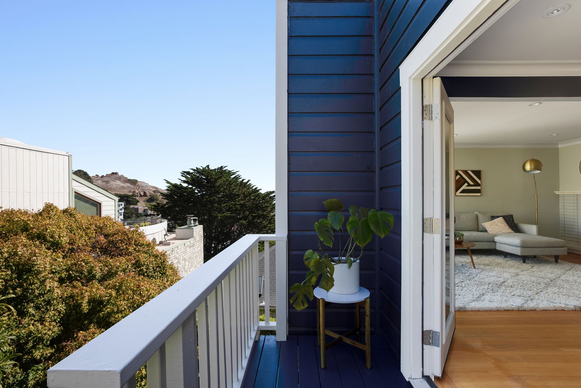View from a private balcony with blue siding and white railings, looking back into the light-filled living room and out toward the lush San Francisco hillside.