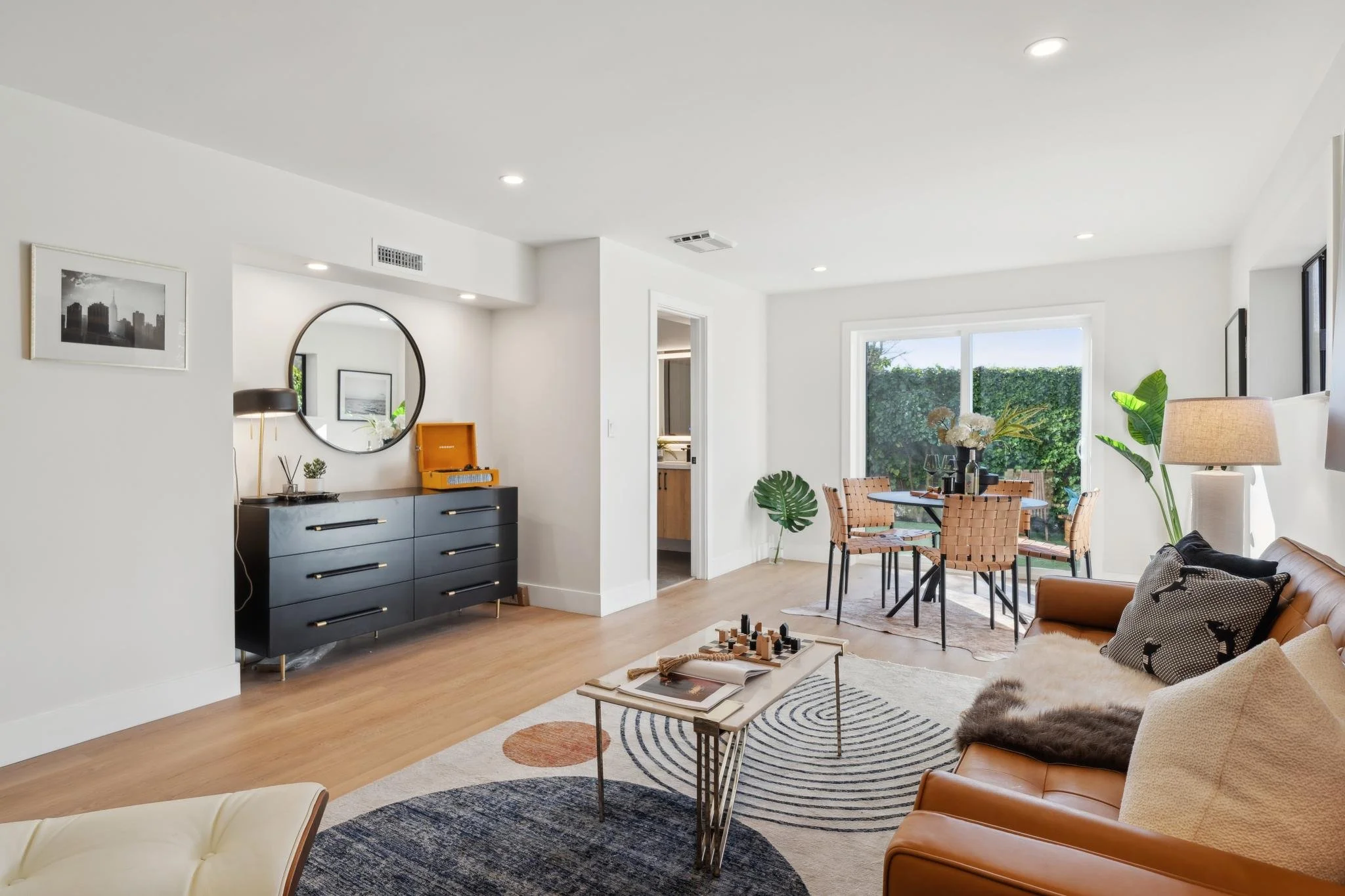 Open-concept lower level lounge with a small dining area, black designer dresser, and large circular mirror, showcasing a seamless indoor-outdoor transition.
