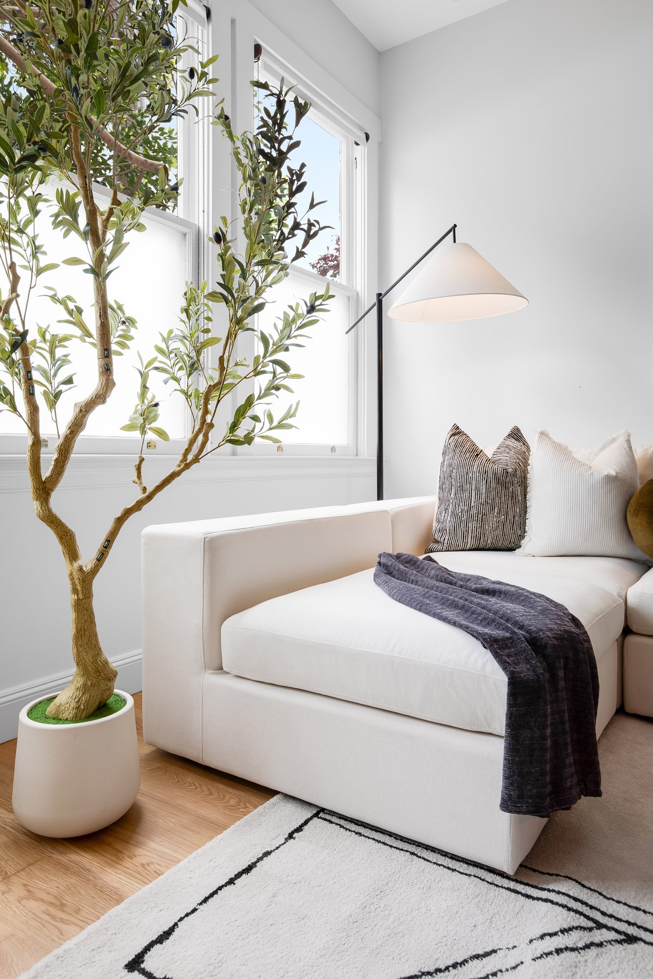 Sunlit living room corner at Castro San Francisco TIC with cream sectional chaise, arc floor lamp, decorative throw pillows, indoor olive tree in white planter, and dual double-hung windows with street views