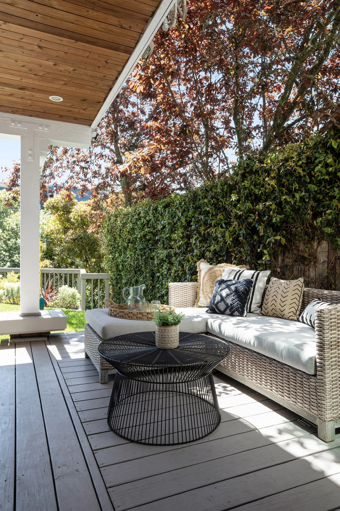 Luxury outdoor lounge area with an L-shaped wicker sofa, black wire coffee table, and a privacy hedge.