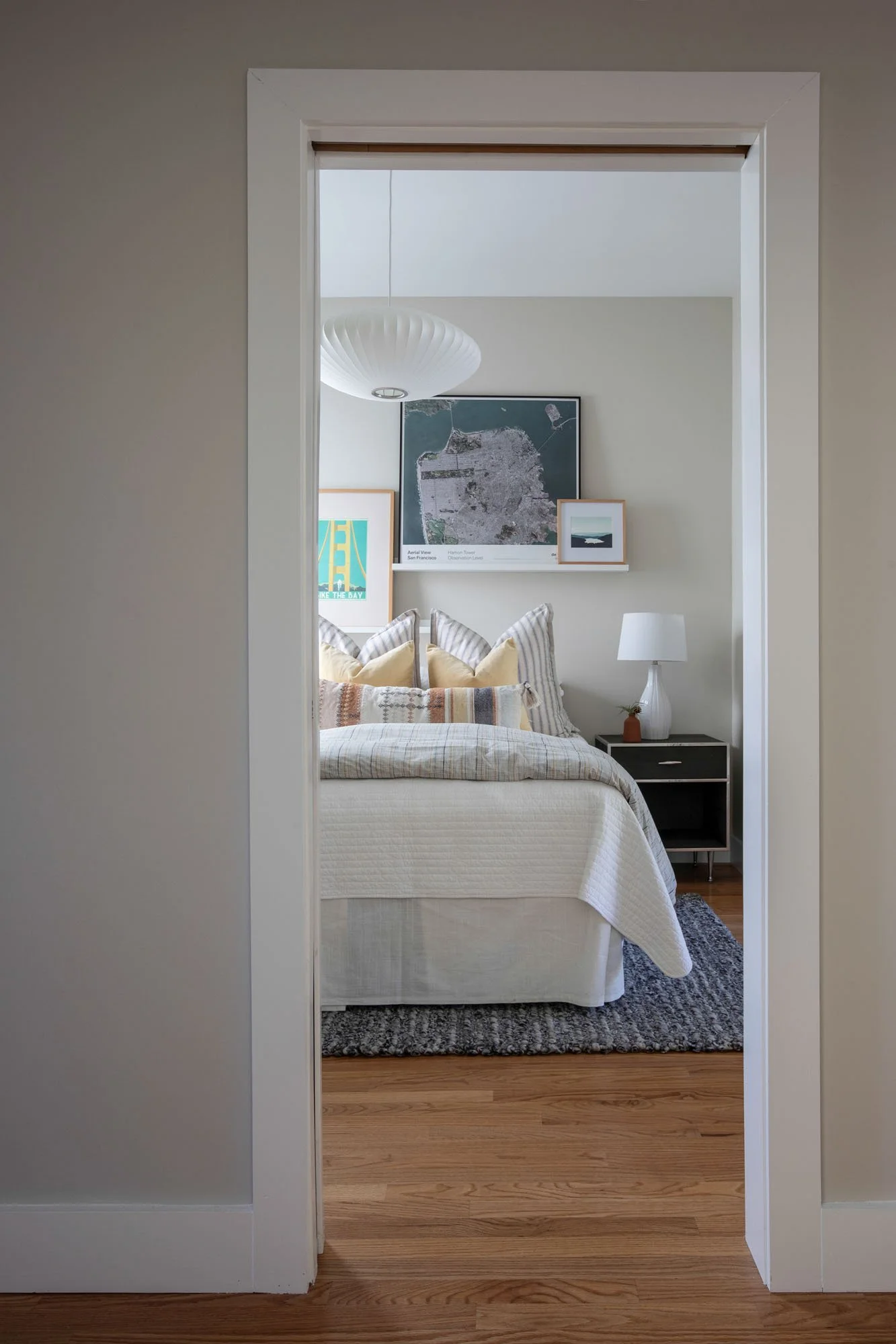 Bright bedroom viewed through a doorway, showcasing a bed with striped pillows, a mid-century modern pendant light, and framed San Francisco maps.
