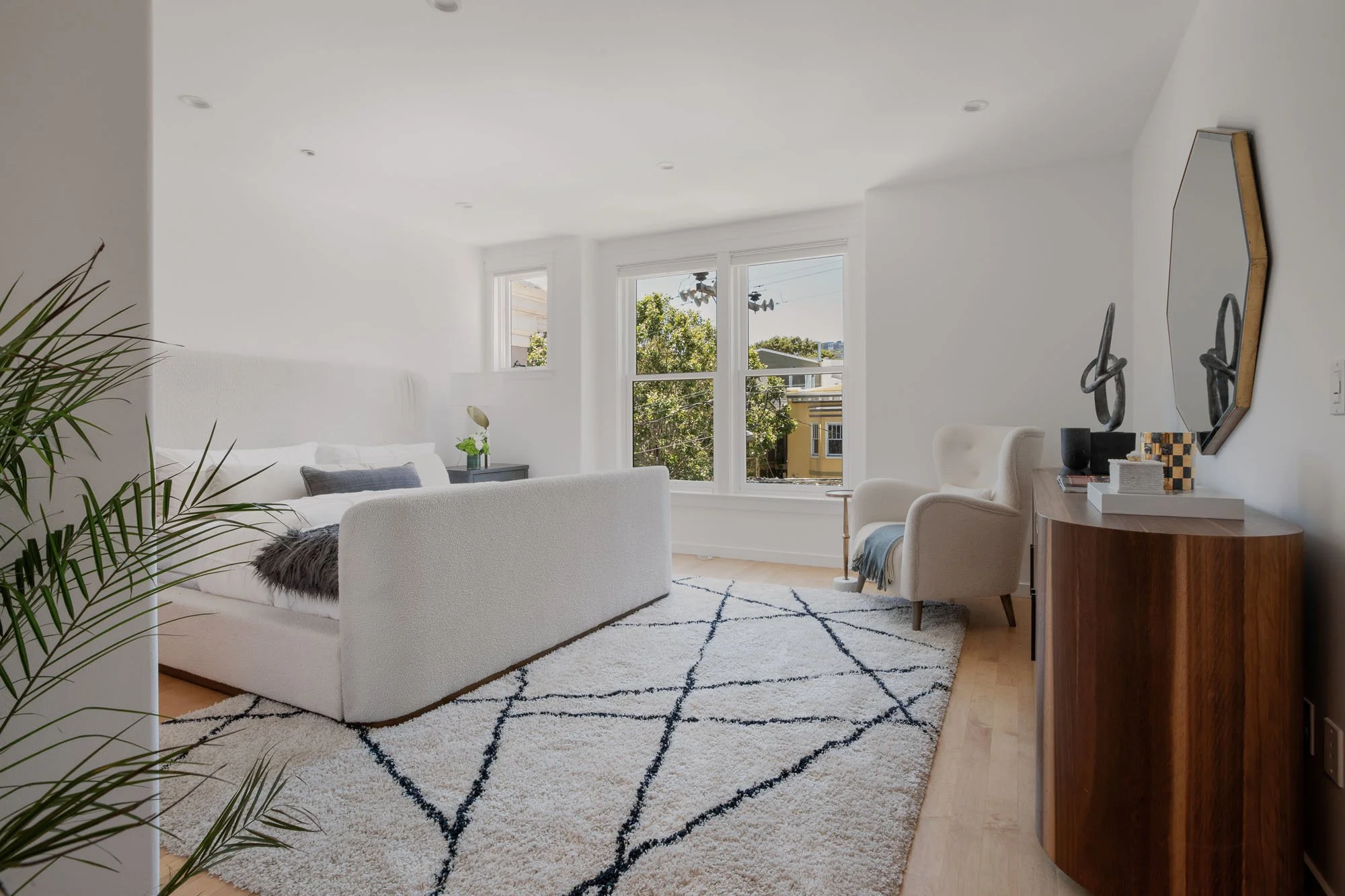 Bright, modern master bedroom with large windows, a white upholstered bed frame, and a minimalist seating area with a mid-century modern wingback chair.