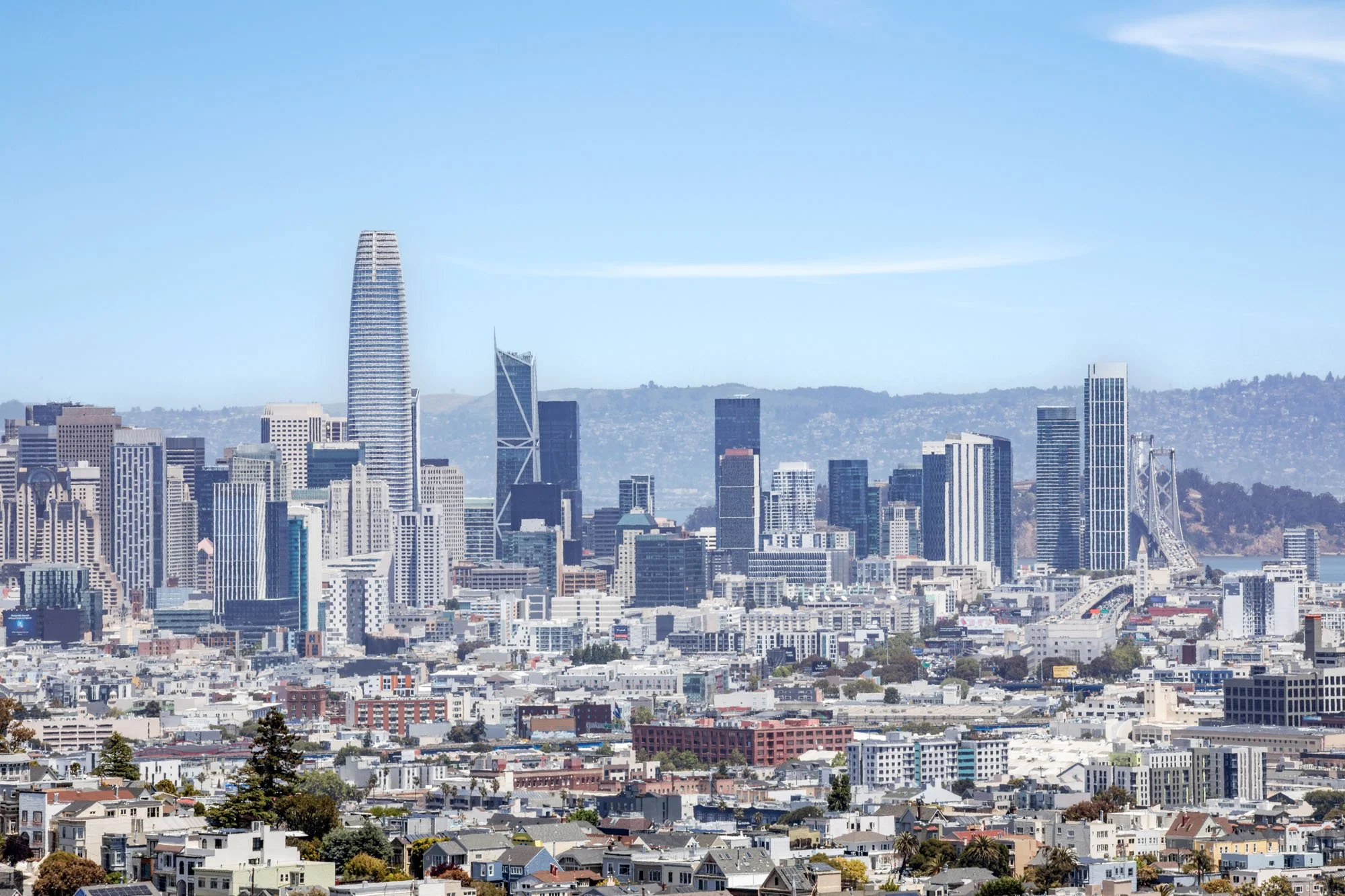 Clear daylight view of downtown San Francisco and the Financial District buildings, capturing the dense urban architecture and the East Bay hills in the distance.