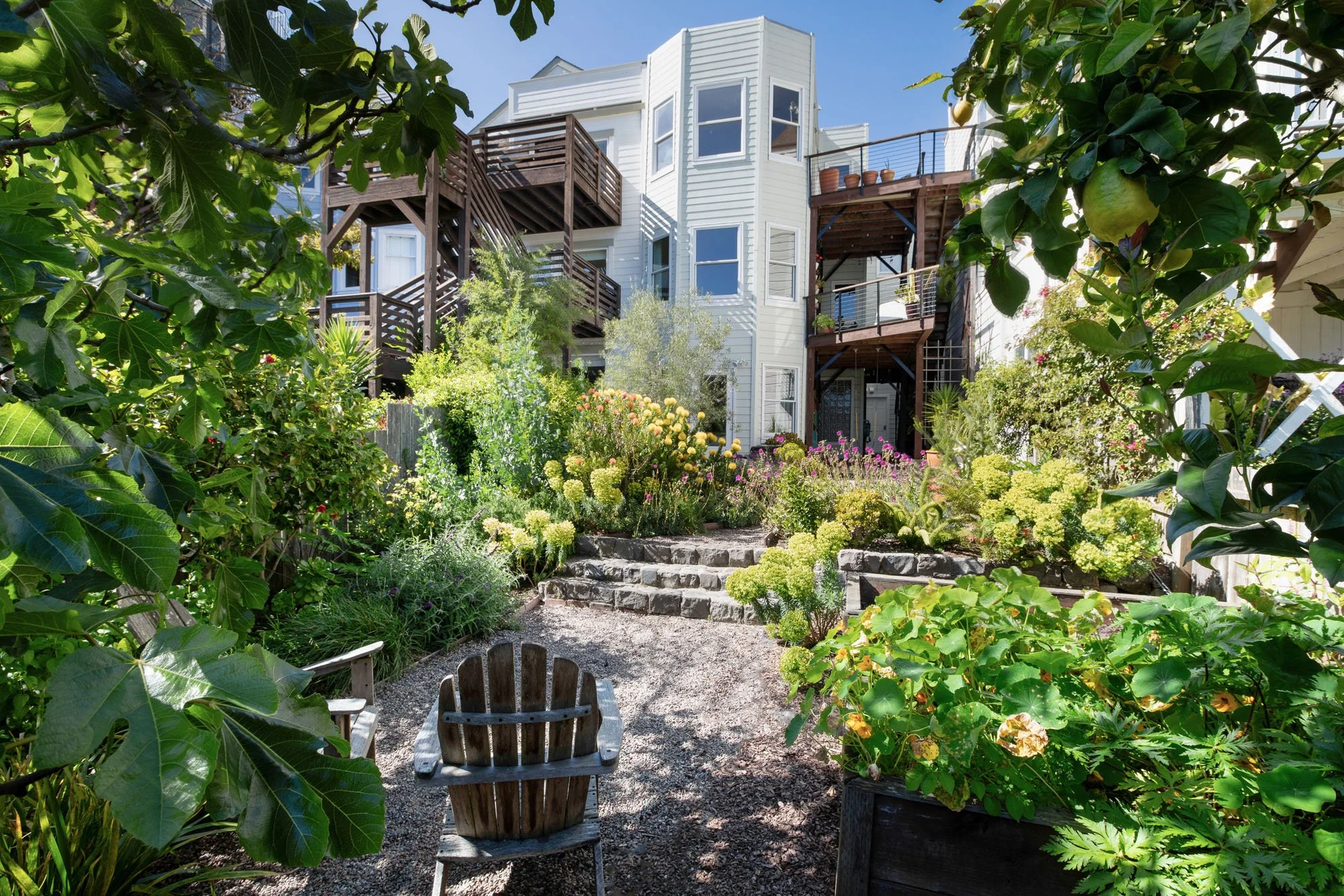 Wide view of a multi-level garden with stone steps, gravel paths, and a variety of blooming plants leading up to a white modern home with wooden decks.