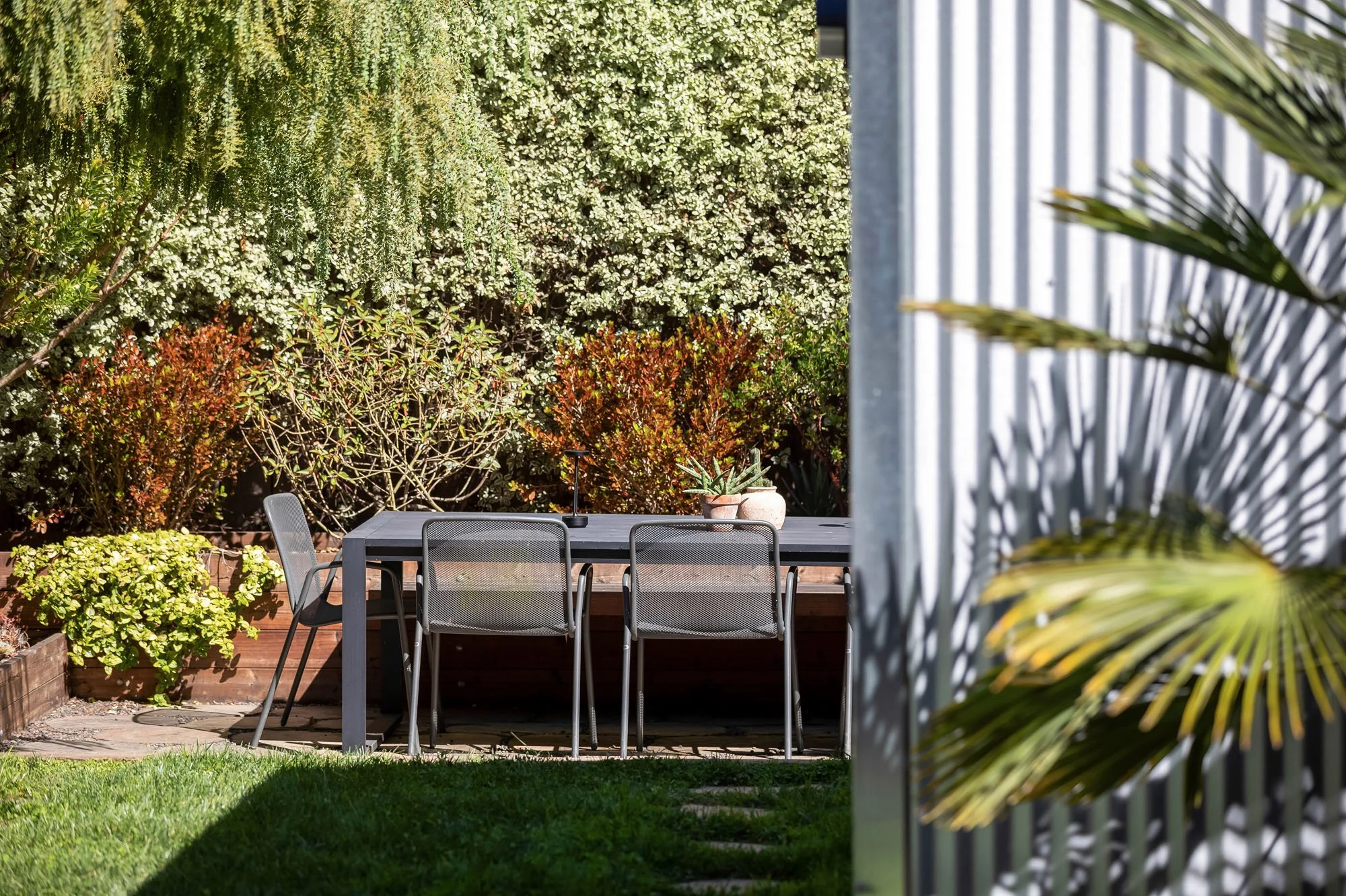 A secluded outdoor dining area with a dark metal table and chairs, set against a dense, private hedge and shaded by mature trees in the 246 Detroit backyard.