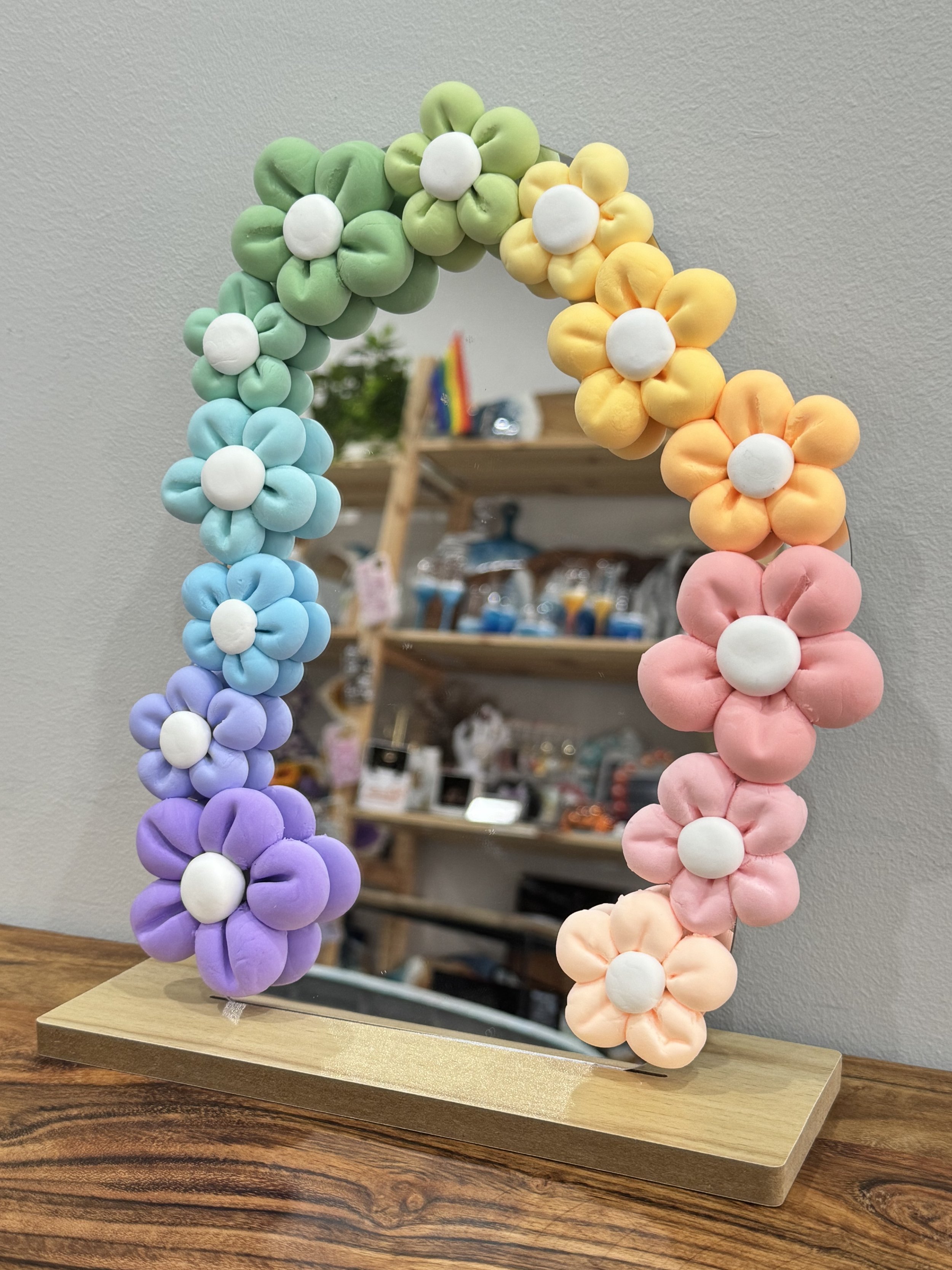 Decorative mirror with colorful plush flowers, arranged in an arch, in a room with wooden shelves and various items in the background.