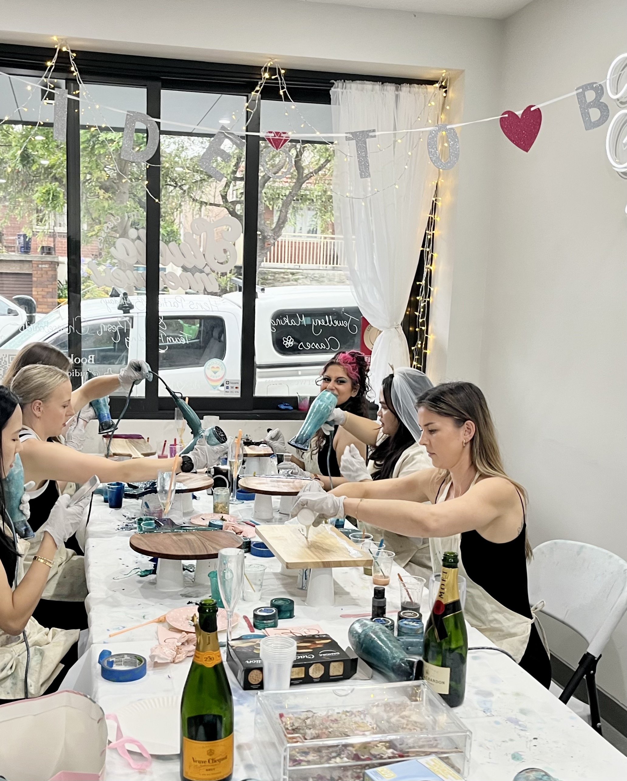 Group of women crafting with paint and tools at a decorated table in a room, champagne bottles visible.