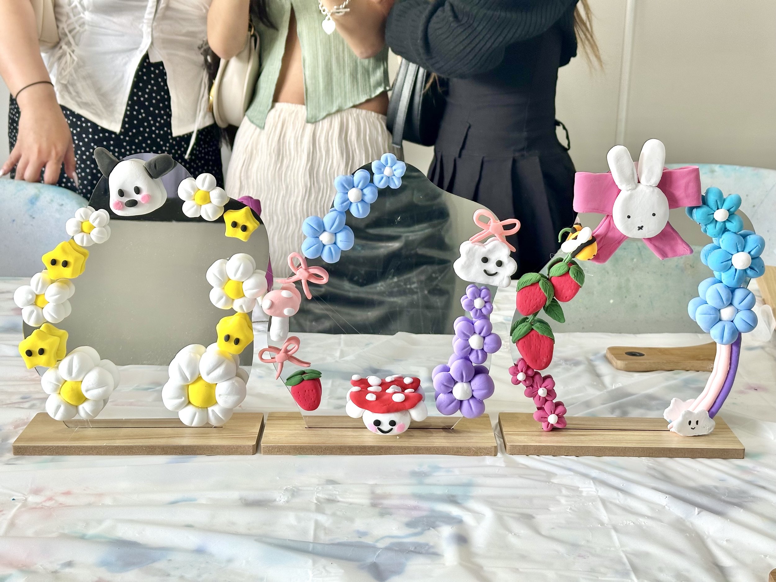 Three decorative mirrors with cute clay figures and flower designs on wooden stands, surrounded by people in background.