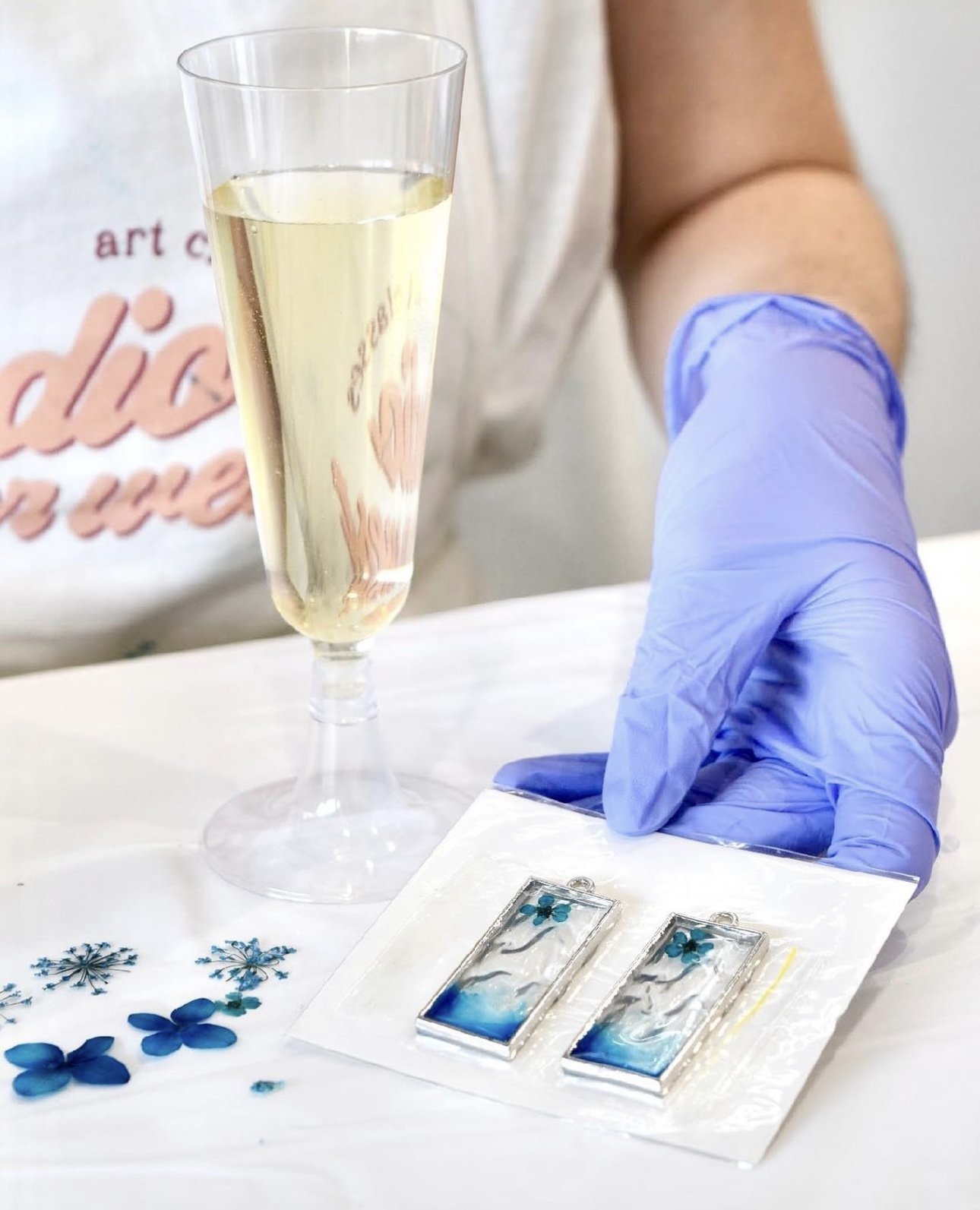 Person wearing a glove holding resin pendants with blue flowers, next to a glass of champagne on a white surface.