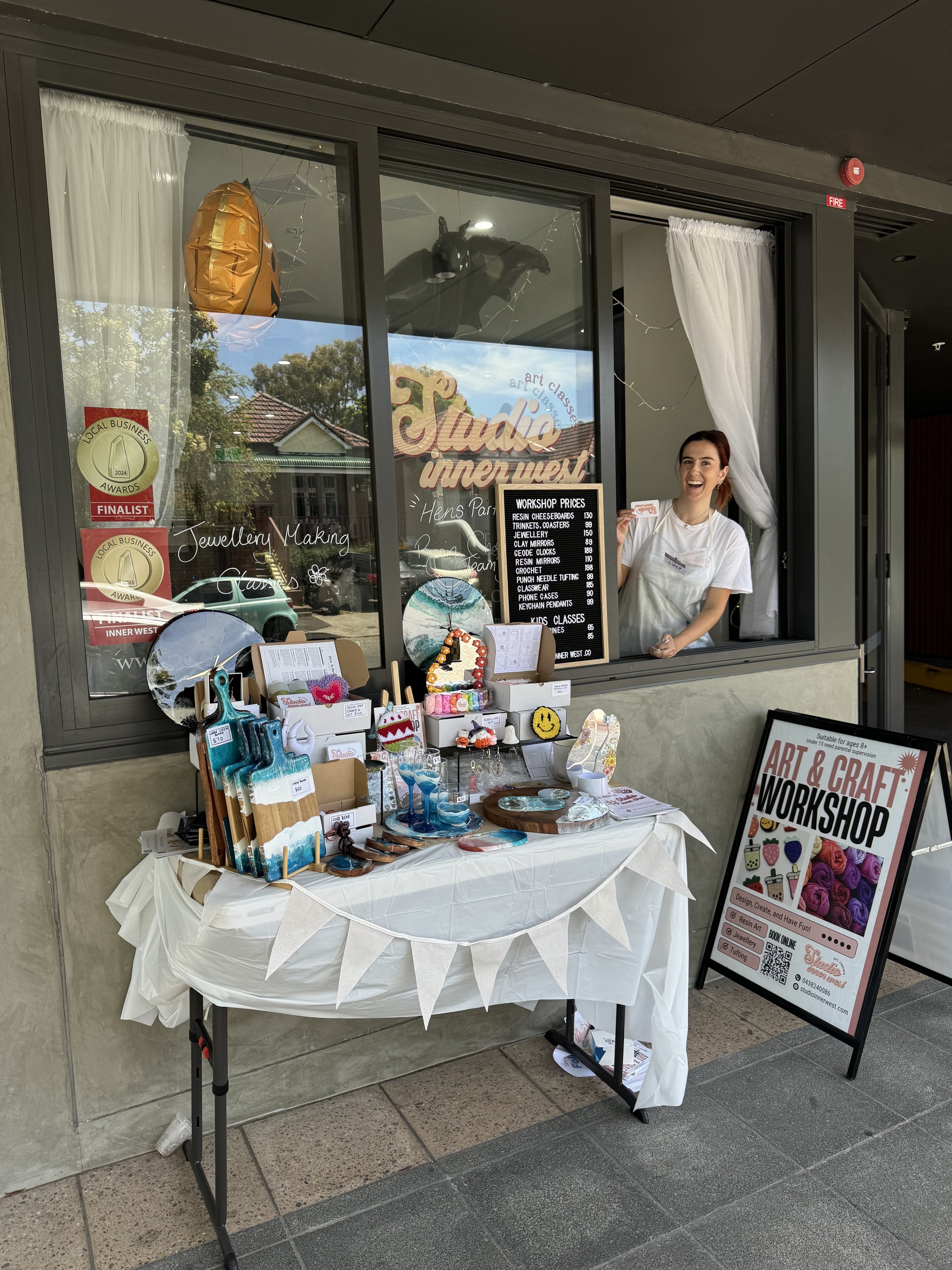 Outdoor display of an art studio with a variety of crafts, including paintings and decorative items, on a table covered with white cloth. A sign promotes an art and craft workshop. A person is smiling through an open window from the studio. The studi