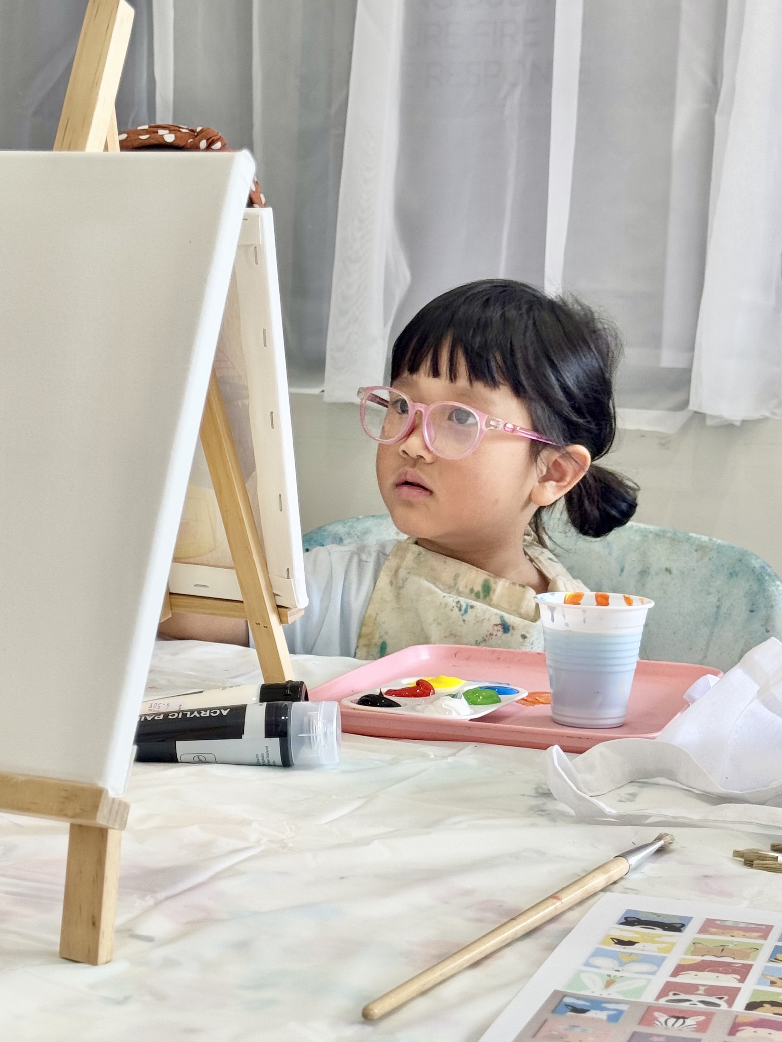 Young child wearing glasses painting at an easel with acrylic paints, paintbrushes, and a paint palette on the table.