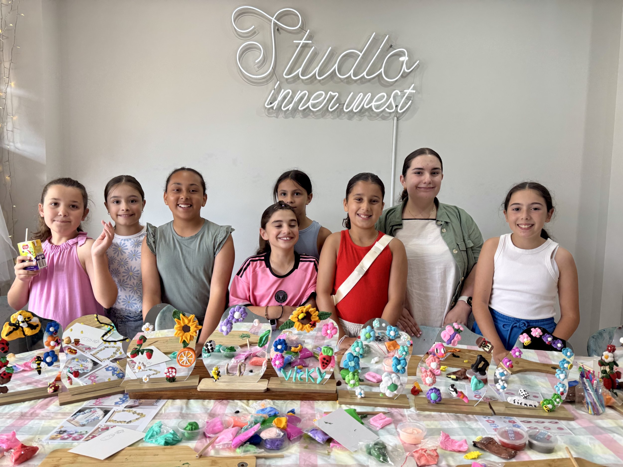 Group of children smiling at a craft table with decorated mirrors and clay art, under a sign that reads "Studio Inner West."