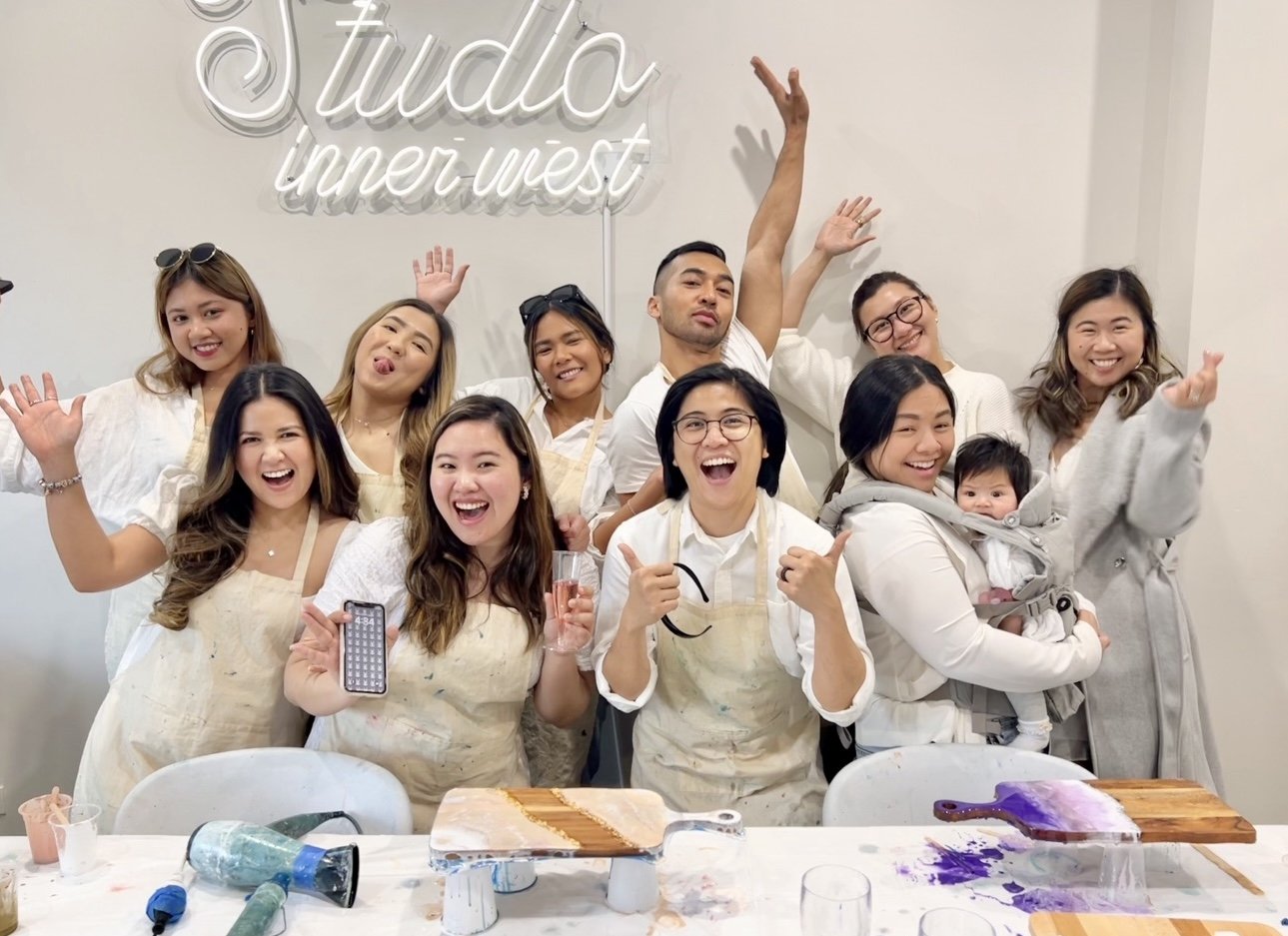 Group of smiling people at Studio Inner West, wearing aprons, some holding art supplies, posing cheerfully indoors.