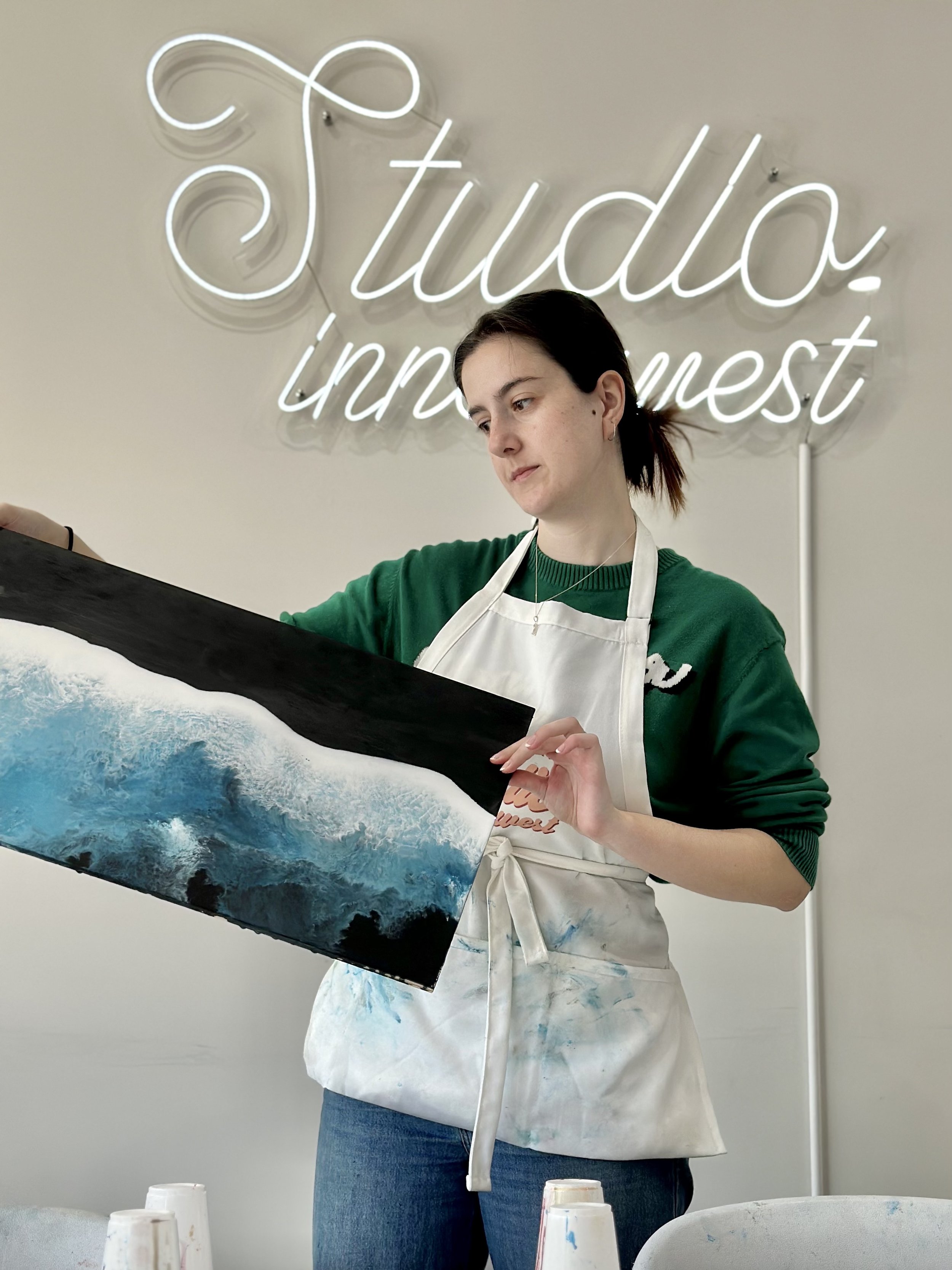 Person holding a painting of ocean waves with a white and blue color palette, standing in front of a neon sign reading "Studio."