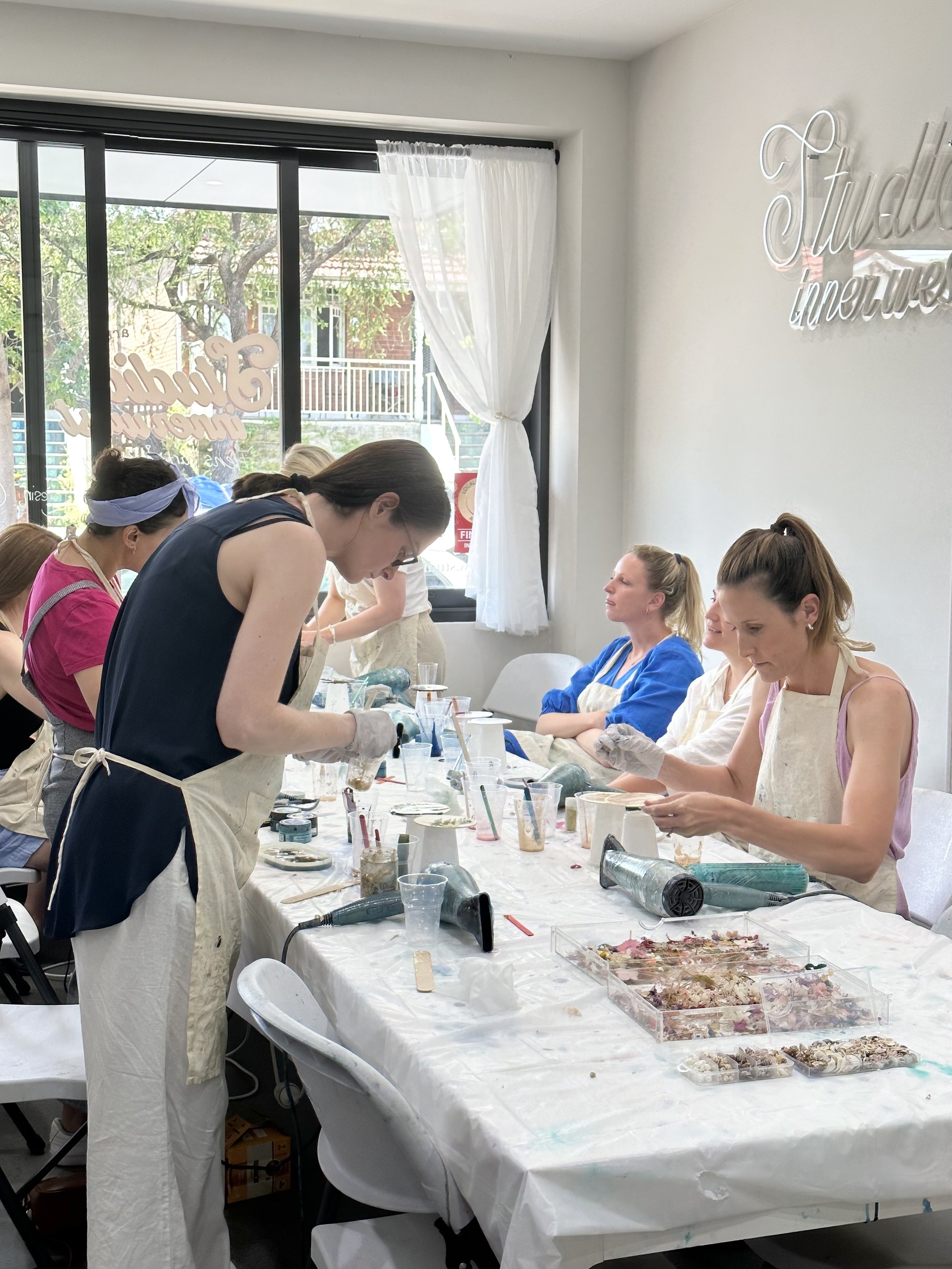 Art class with participants creating crafts on a table, using materials like shells and paint, in a sunlit studio.