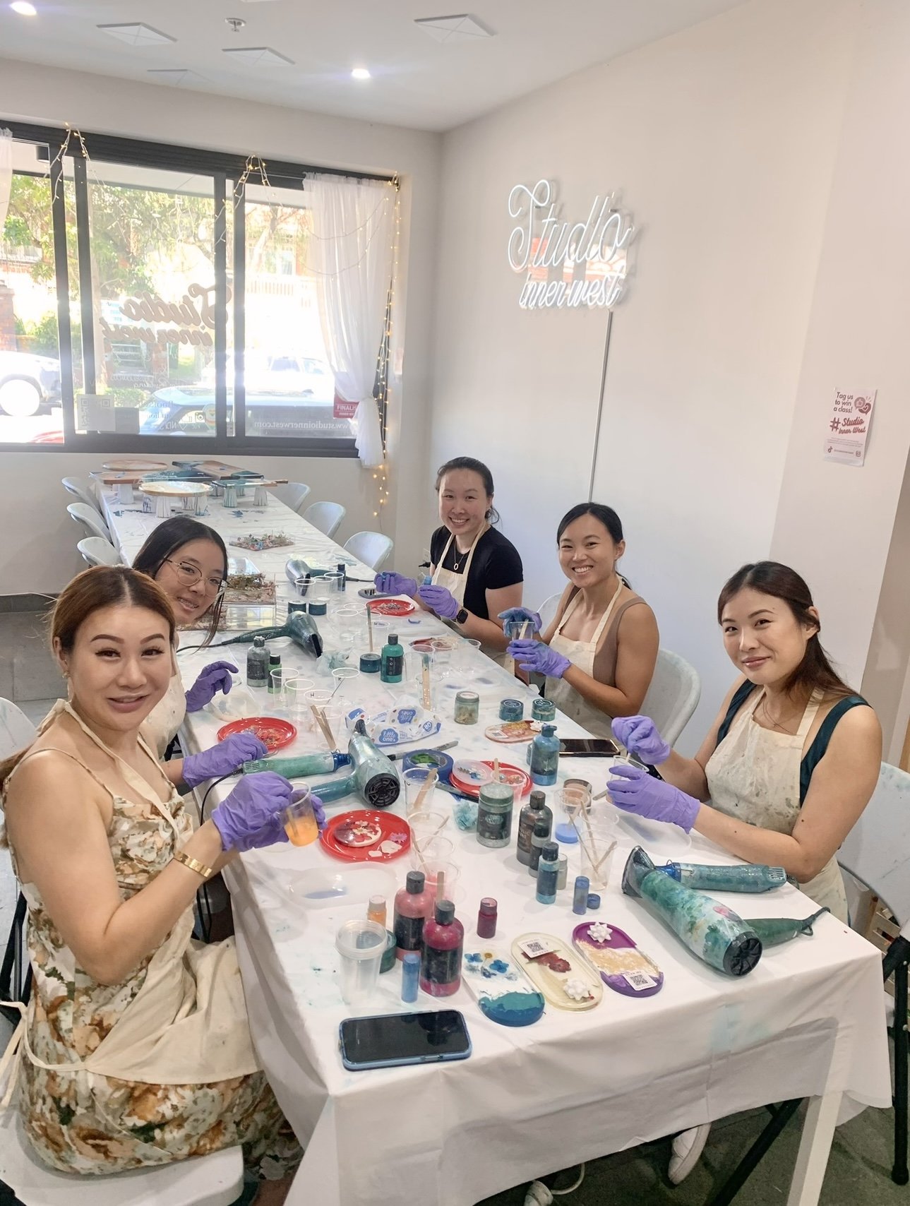 Group of women at a table in an art studio, wearing aprons and gloves, engaged in a craft activity involving paints and hair dryers.