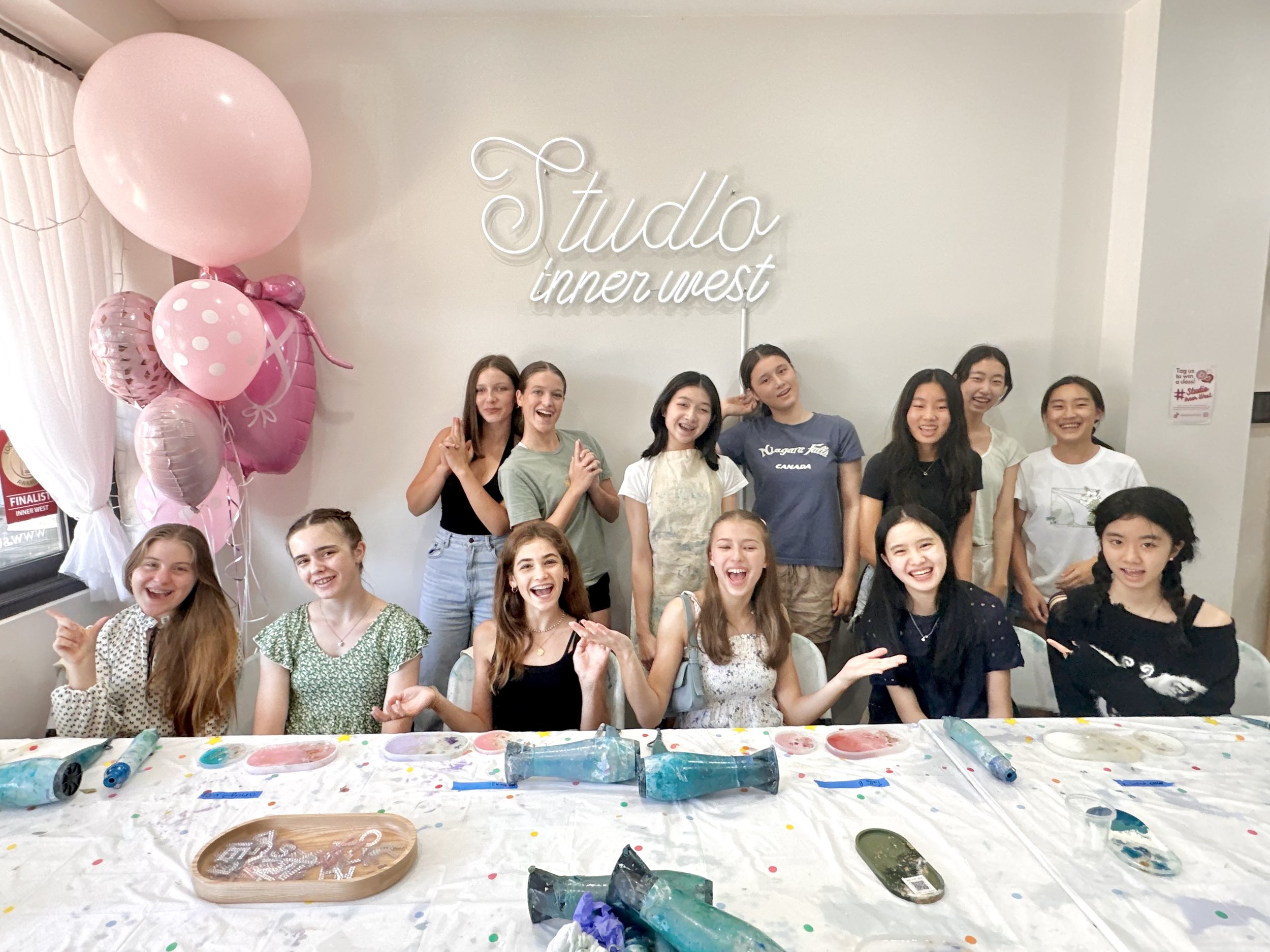 A group of girls smiling in front of a sign that reads 'Studio Inner West'. They are surrounded by crafting materials on a table, with pink balloons in the background, suggesting a fun group activity or workshop.