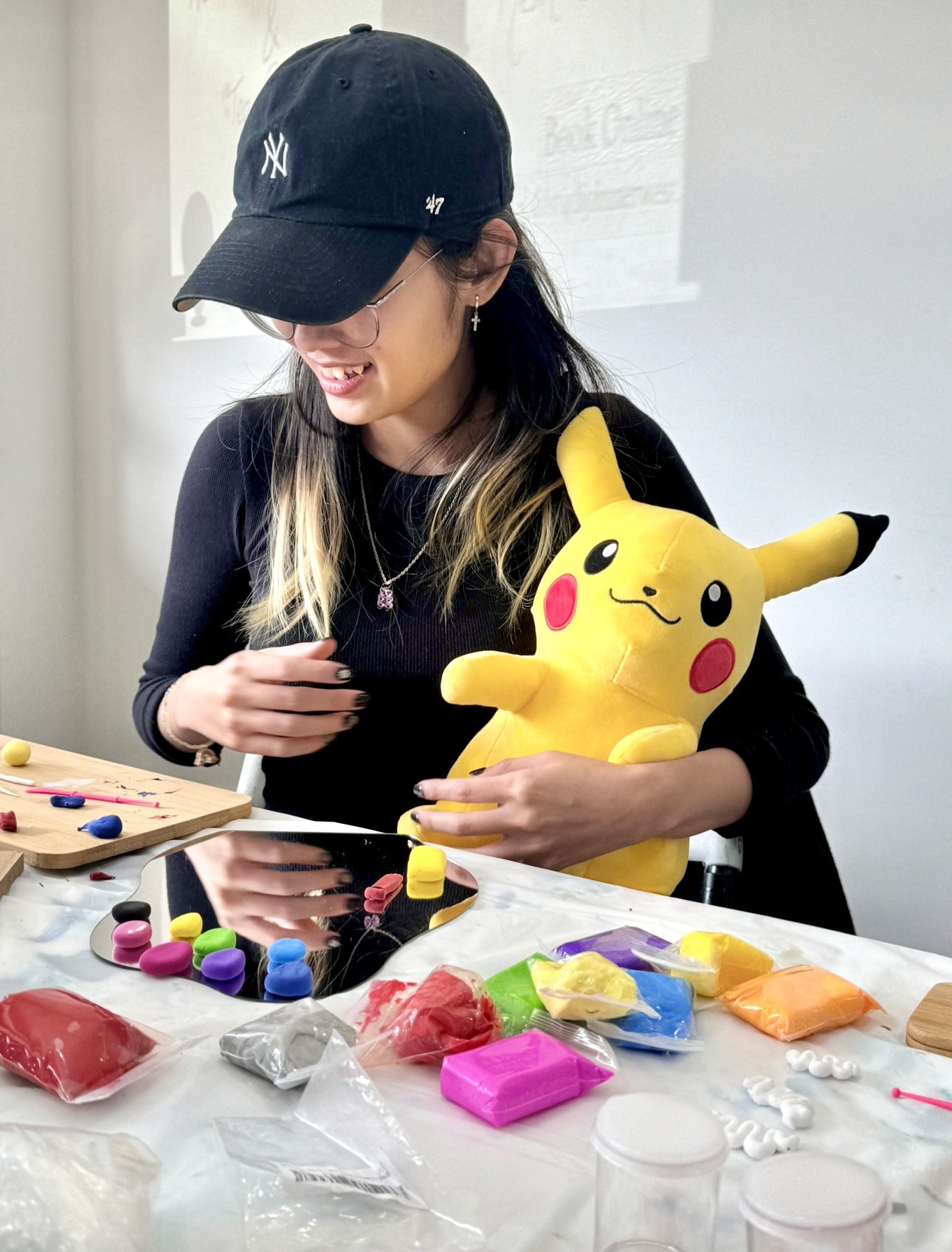 Person wearing a black cap holding a Pikachu plush toy, seated at a table with colorful crafting materials like clay and paint.